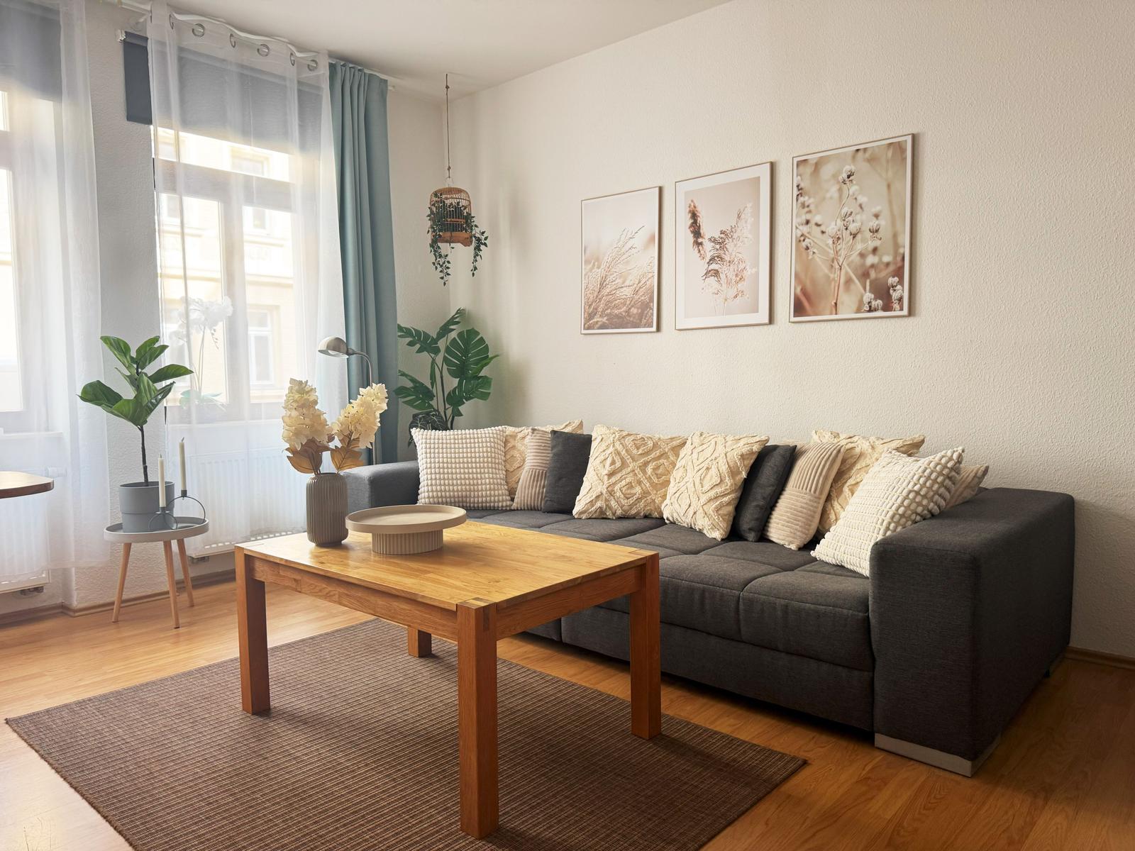 Ein Wohnzimmer mit grauem Sofa, Holztisch und Pflanzen. An der Wand hängen drei Bilder.