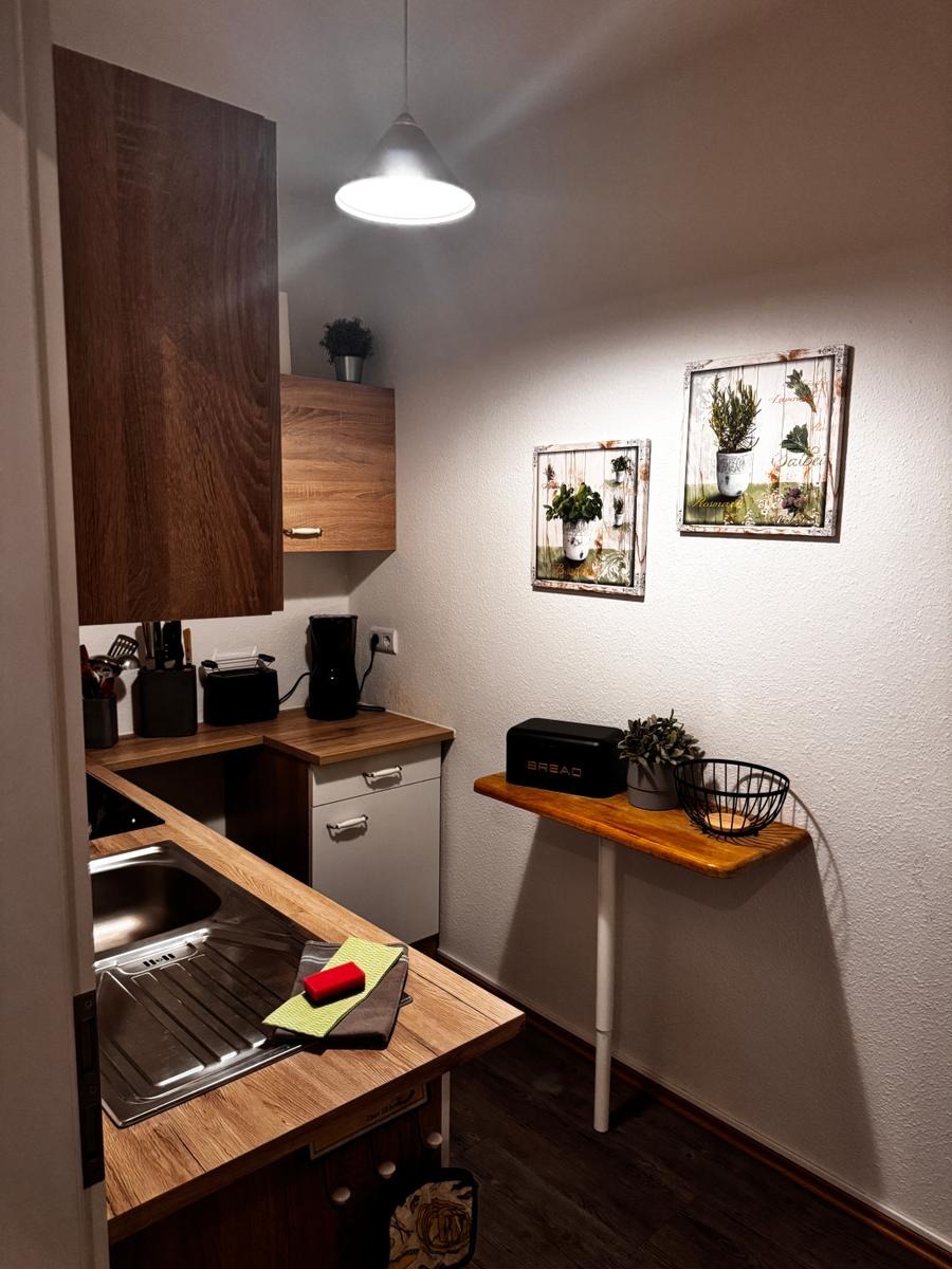 Küche mit Holzoberflächen, Spüle und Wandbildern. Nebenraum mit kleinem Tisch und Pflanzen.