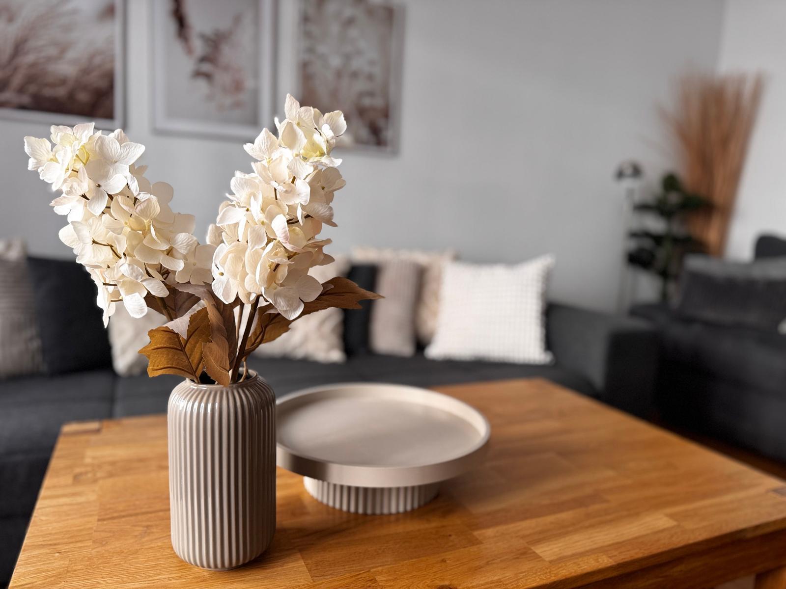 Ein Wohnzimmer mit Holztisch, Vase mit weißen Blumen und Sofa im Hintergrund.