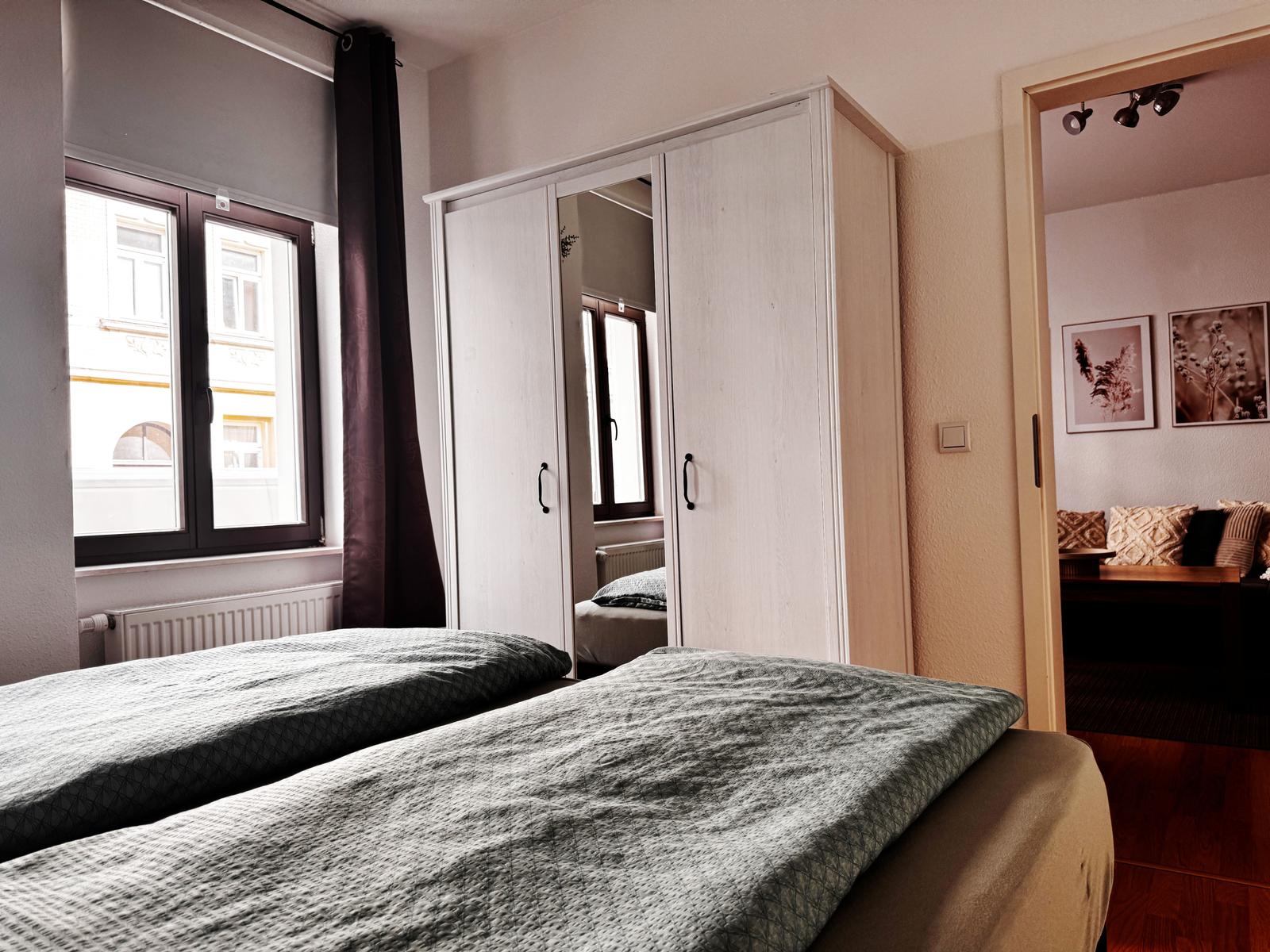 Zimmer mit Doppelbett, Kleiderschrank und Fenster. Durchgang zu Wohnbereich mit Sofa.