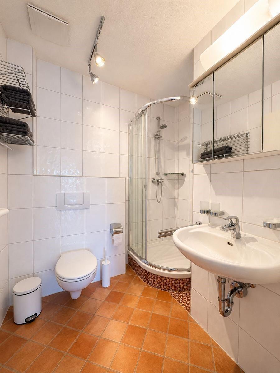 Badezimmer mit Dusche, Waschbecken und Toilette. Fliesenboden und Spiegelschrank.