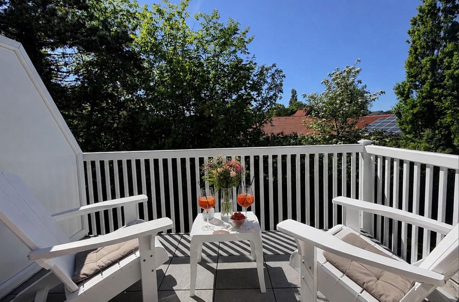 Terrasse mit zwei weißen Stühlen, Tisch und Getränken. Blick auf Bäume und Dächer.