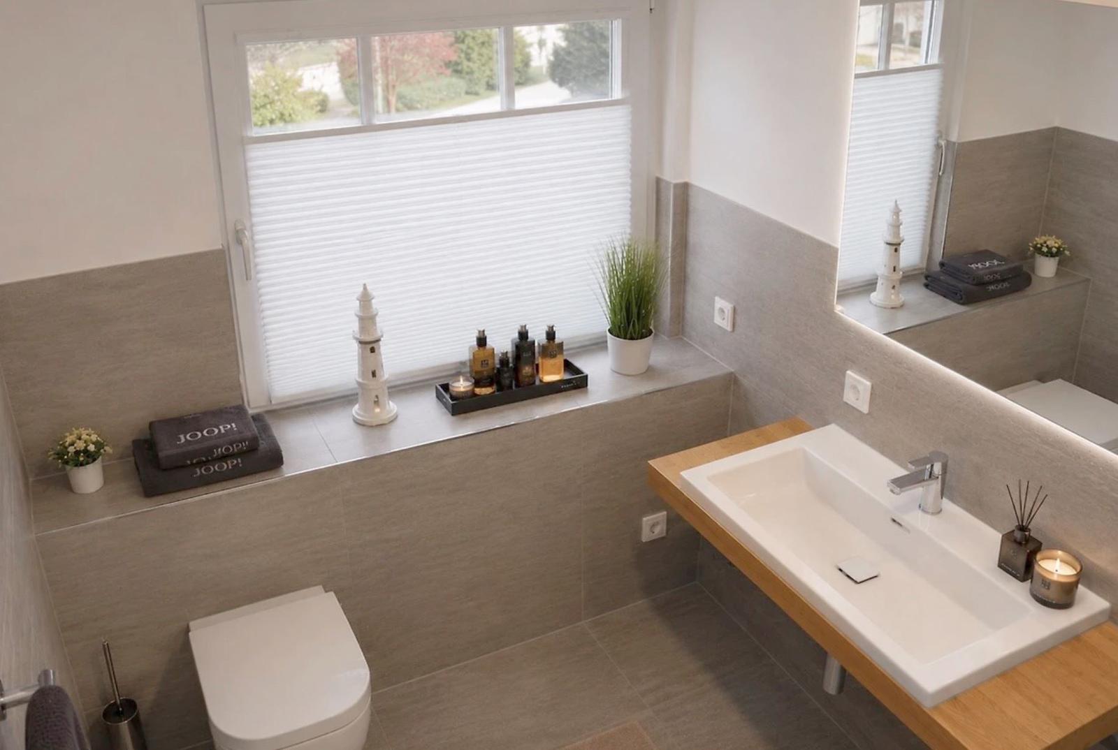 Badezimmer mit Fenster, Waschbecken und Toilette. Auf dem Fensterbrett stehen Dekorationsgegenstände.