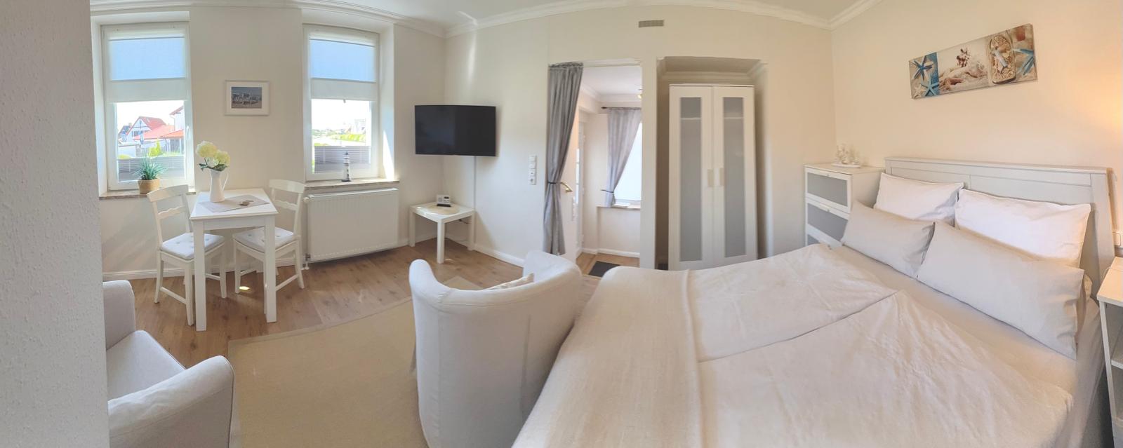 Schlafzimmer mit Bett, Schrank und TV. Fenster mit Tisch und Stühlen. Wandbild über dem Bett.