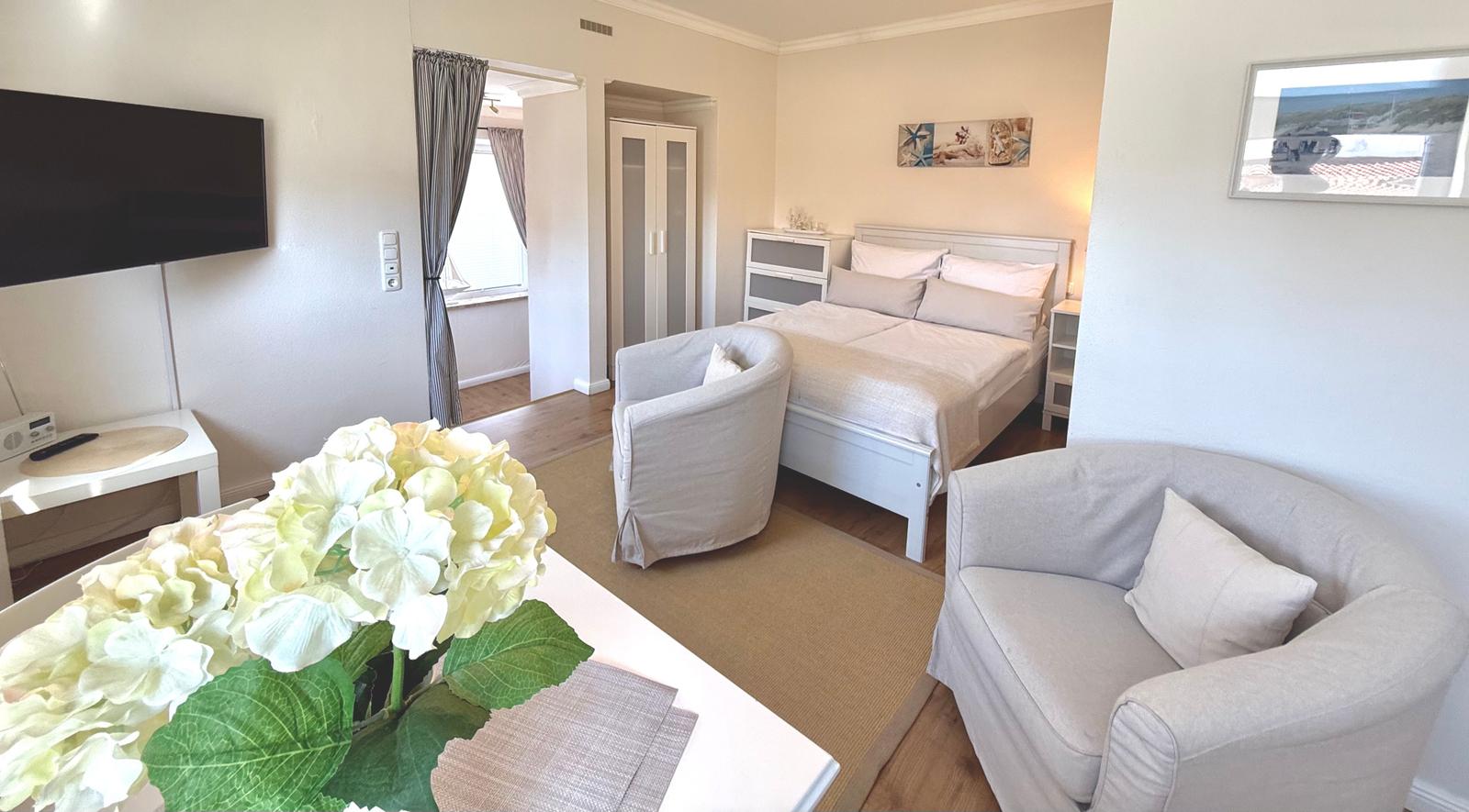 Ein Schlafzimmer mit Bett, zwei Sesseln, Fernseher und Blumenarrangement.