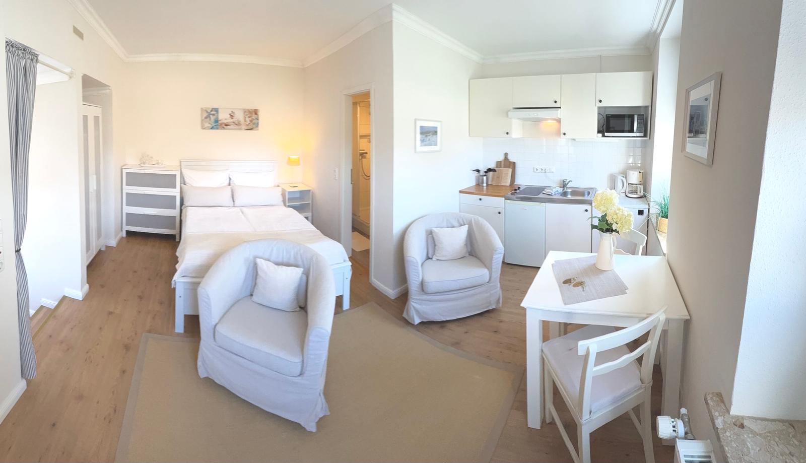Schlafzimmer mit Bett, zwei Sesseln, Küchenbereich und Esstisch.
