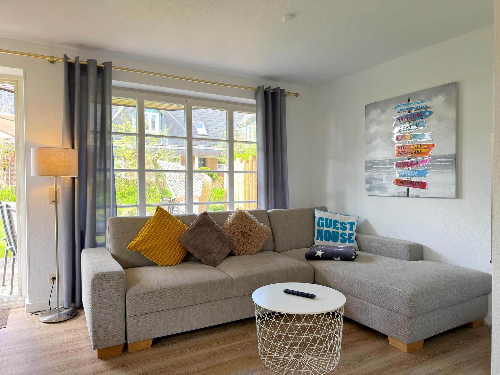Ein Wohnzimmer mit grauem Sofa, Kissen und einem runden Tisch. An der Wand hängt ein Bild mit Wegweisern.