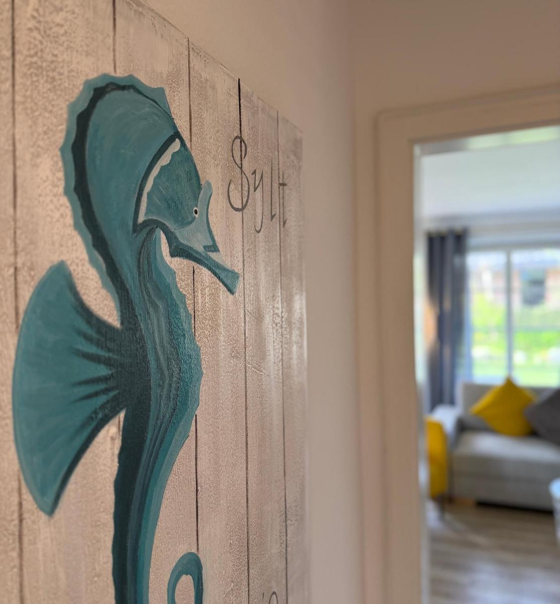 Holzpaneel mit Seepferdchenmalerei und Schriftzug Sylt an Wand. Im Hintergrund ist Wohnzimmer sichtbar.