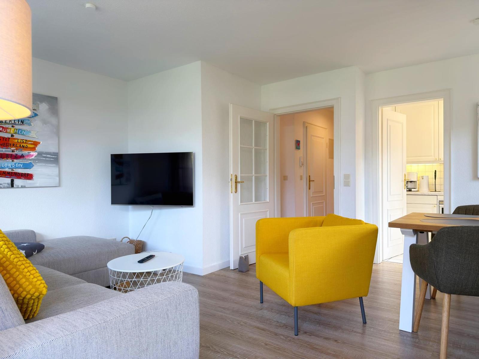Wohnzimmer mit gelbem Sessel, Sofa und Fernseher. Küche und Flur sind sichtbar.