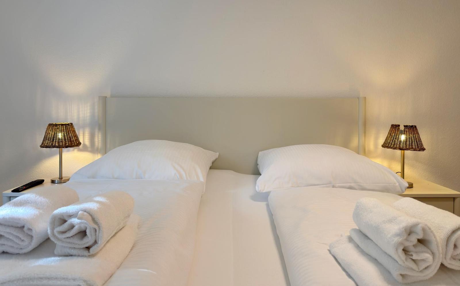 Zwei Betten mit weißen Laken und Handtüchern, jeweils mit Nachttisch und Lampe.
