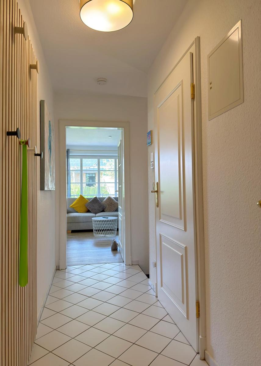 Flur mit weißen Fliesen führt in ein Wohnzimmer mit Sofa und Fenster.