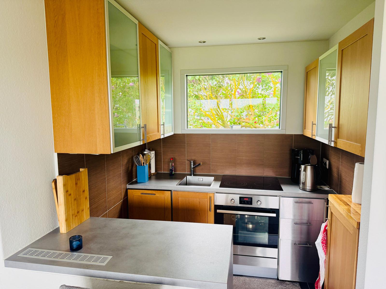 Küche mit Holzschränken, Edelstahlgeräten und Fenster zur grünen Außenseite.