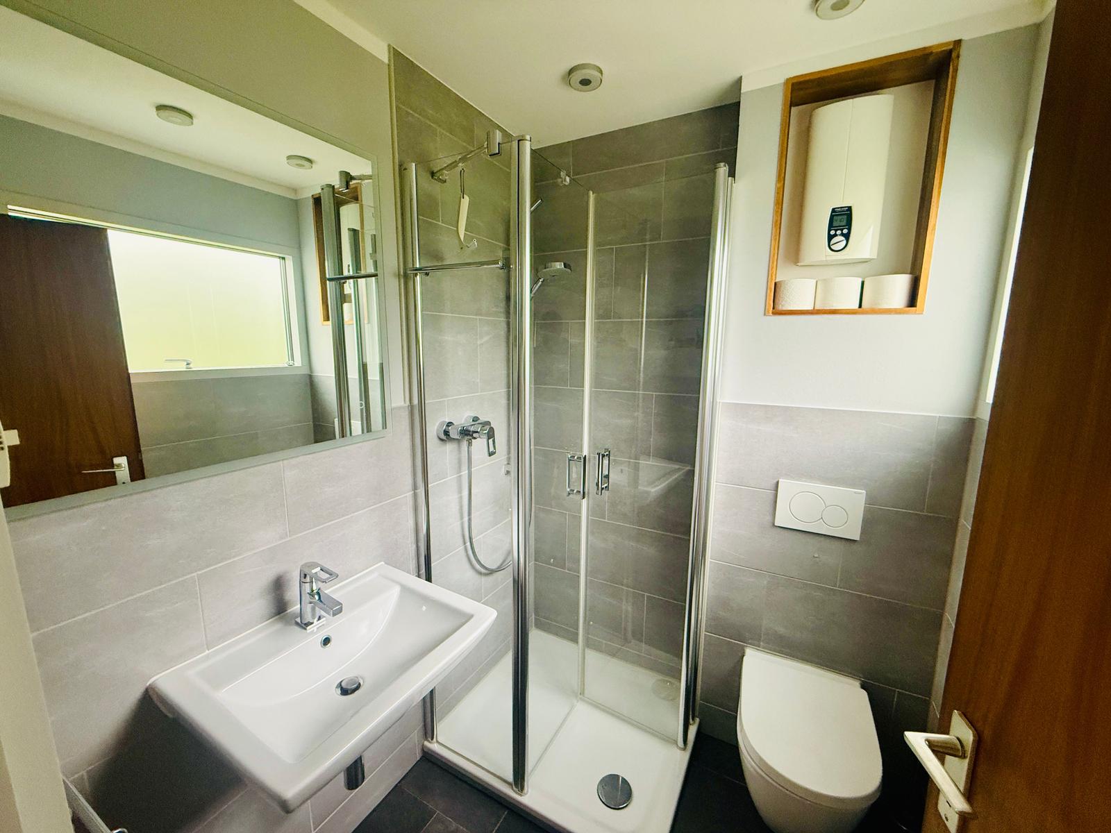 Badezimmer mit Dusche, Waschbecken und Toilette. Graue Fliesen und Spiegel.