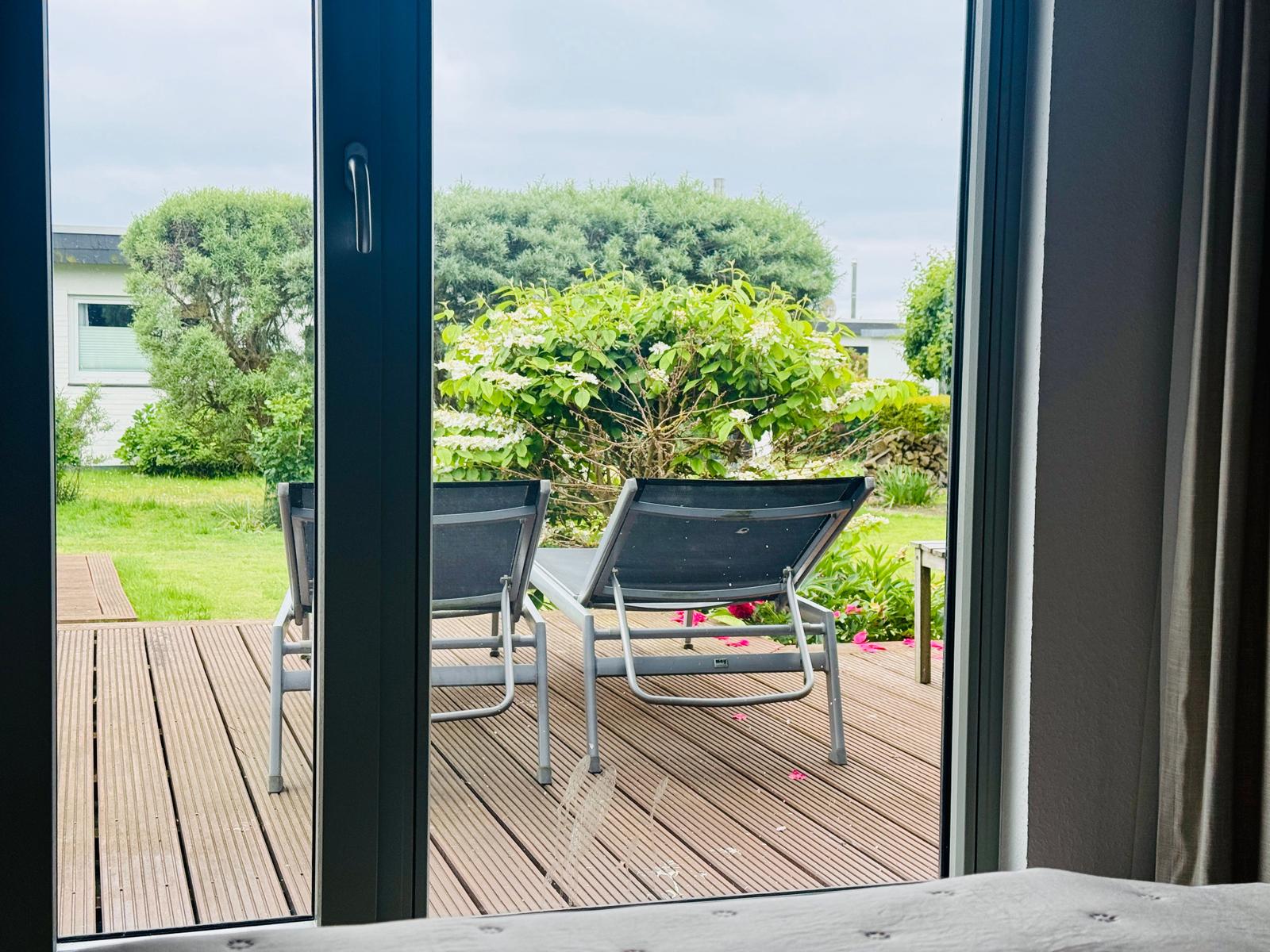 Durch die Glastür blickt man auf eine Terrasse mit zwei Liegestühlen und einen grünen Garten.