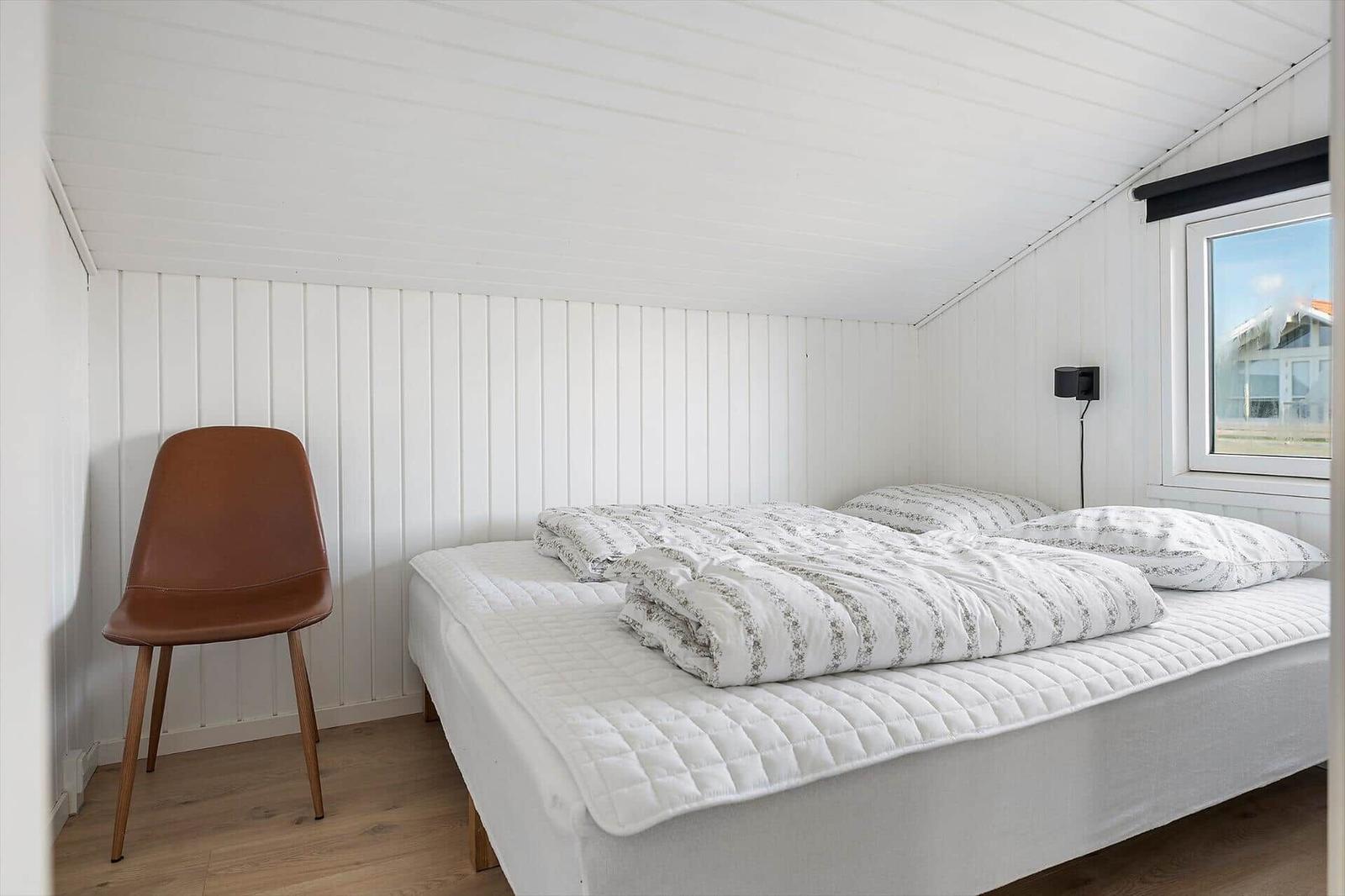 Bedroom with bed, brown chair, and window. White walls and wooden floor.