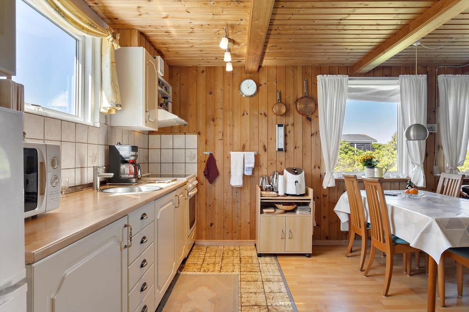 Kitchen with dining area, wooden walls, and windows. Kitchen counter, table, chairs, appliances.