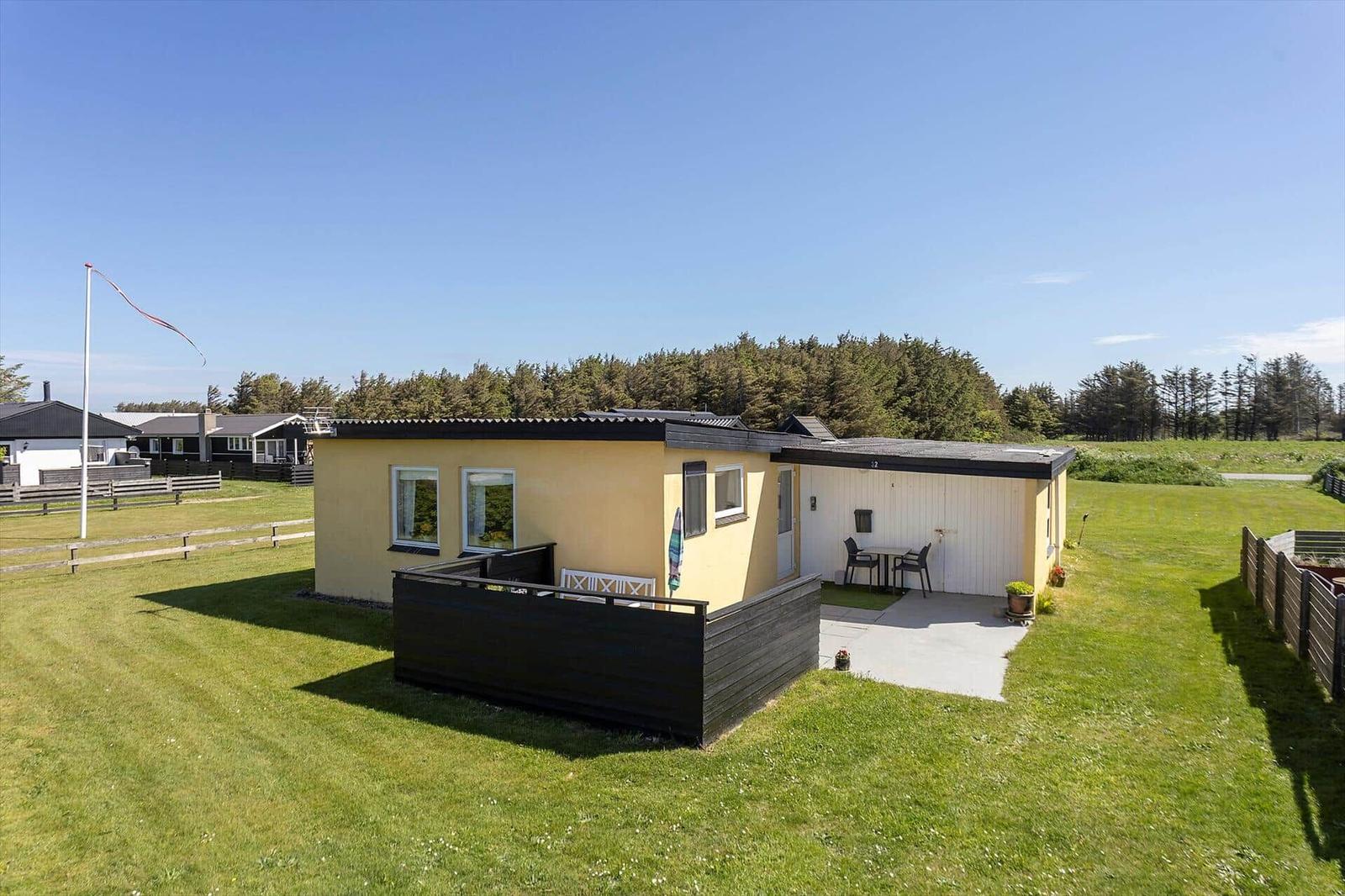 A yellow holiday home with terrace and garden. Trees and a fence are visible in the background.
