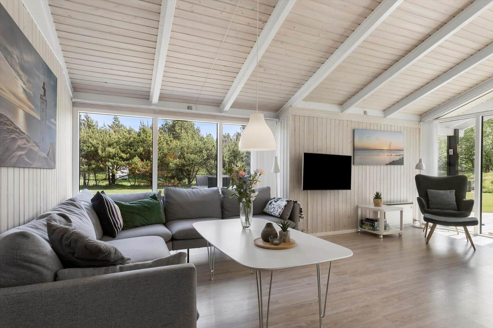 Living room with sofa, table, and TV. Large windows overlooking green area.