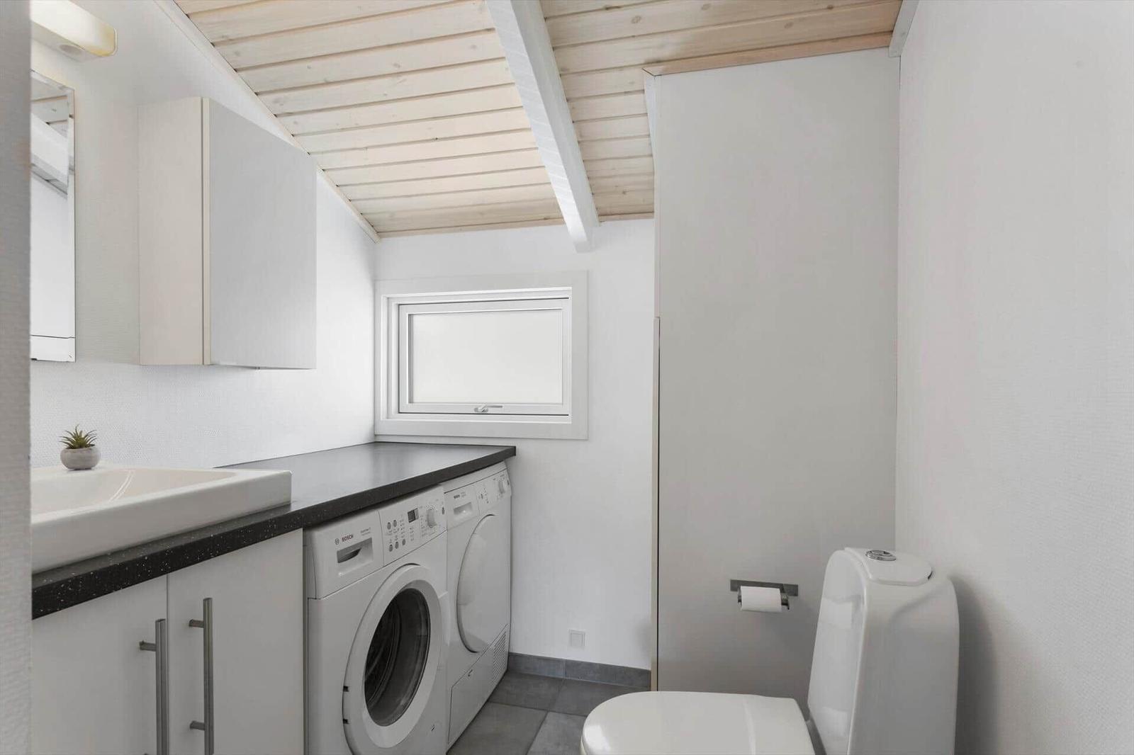 Washroom with washing machine, sink, and toilet under wooden ceiling.