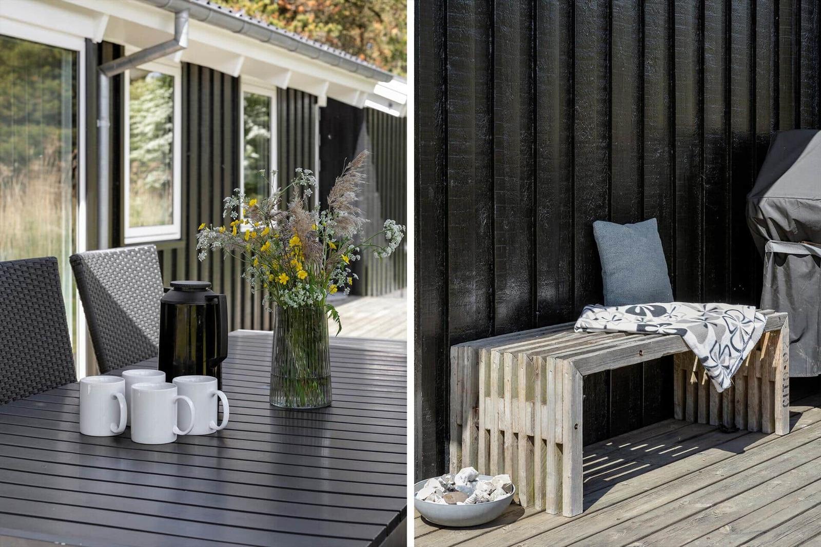 Terrace with table, chairs, flowers, and coffee mugs. Wooden bench with cushion and blanket against black wall.