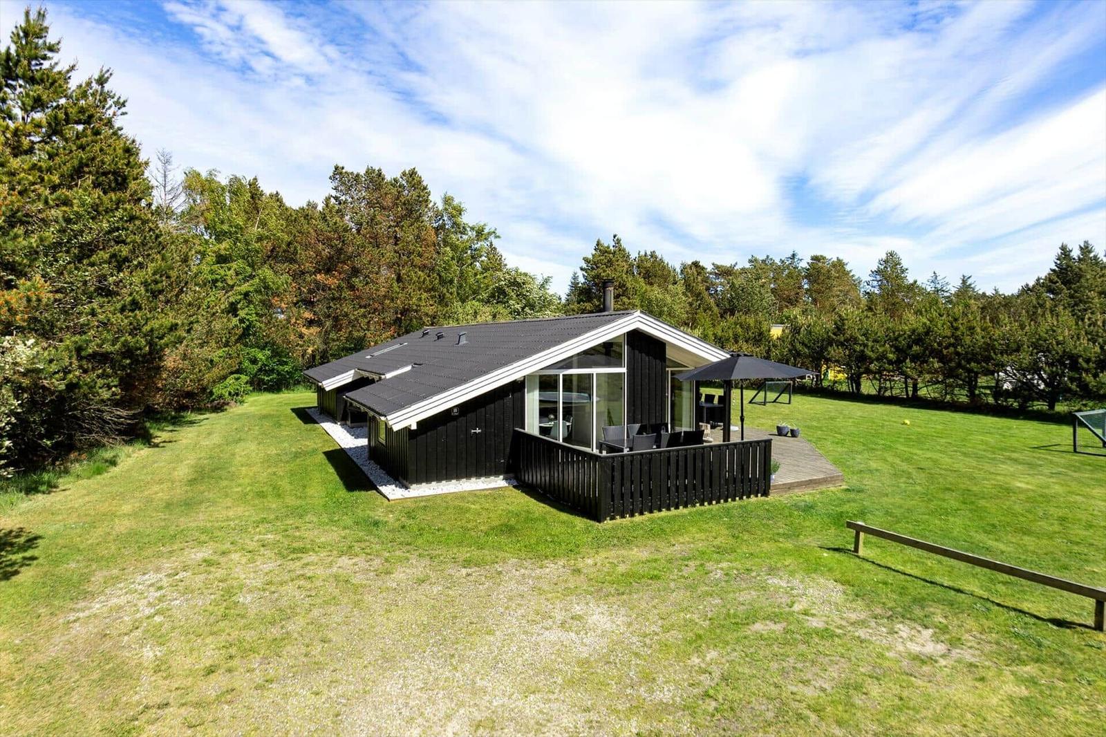 Black wooden house with terrace and garden. Surrounded by trees under blue sky.
