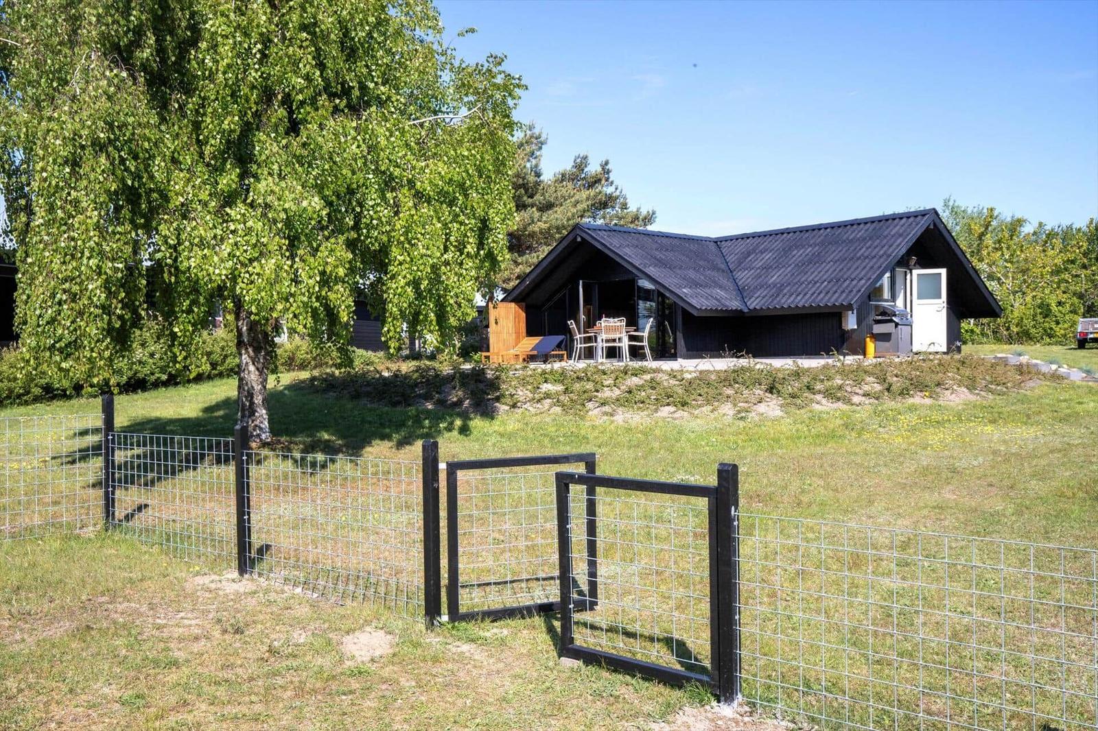 A house with a dark roof and terrace stands on a green lawn behind a fence.