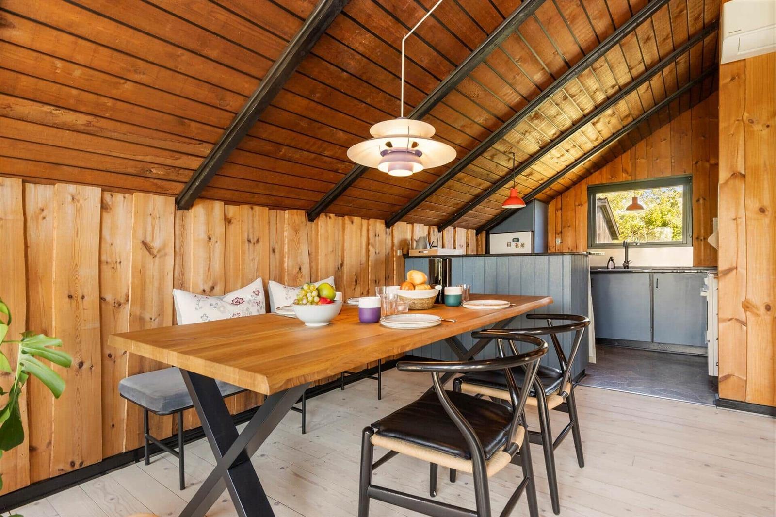 Dining area with wooden table, chairs, and kitchen under wooden ceiling.