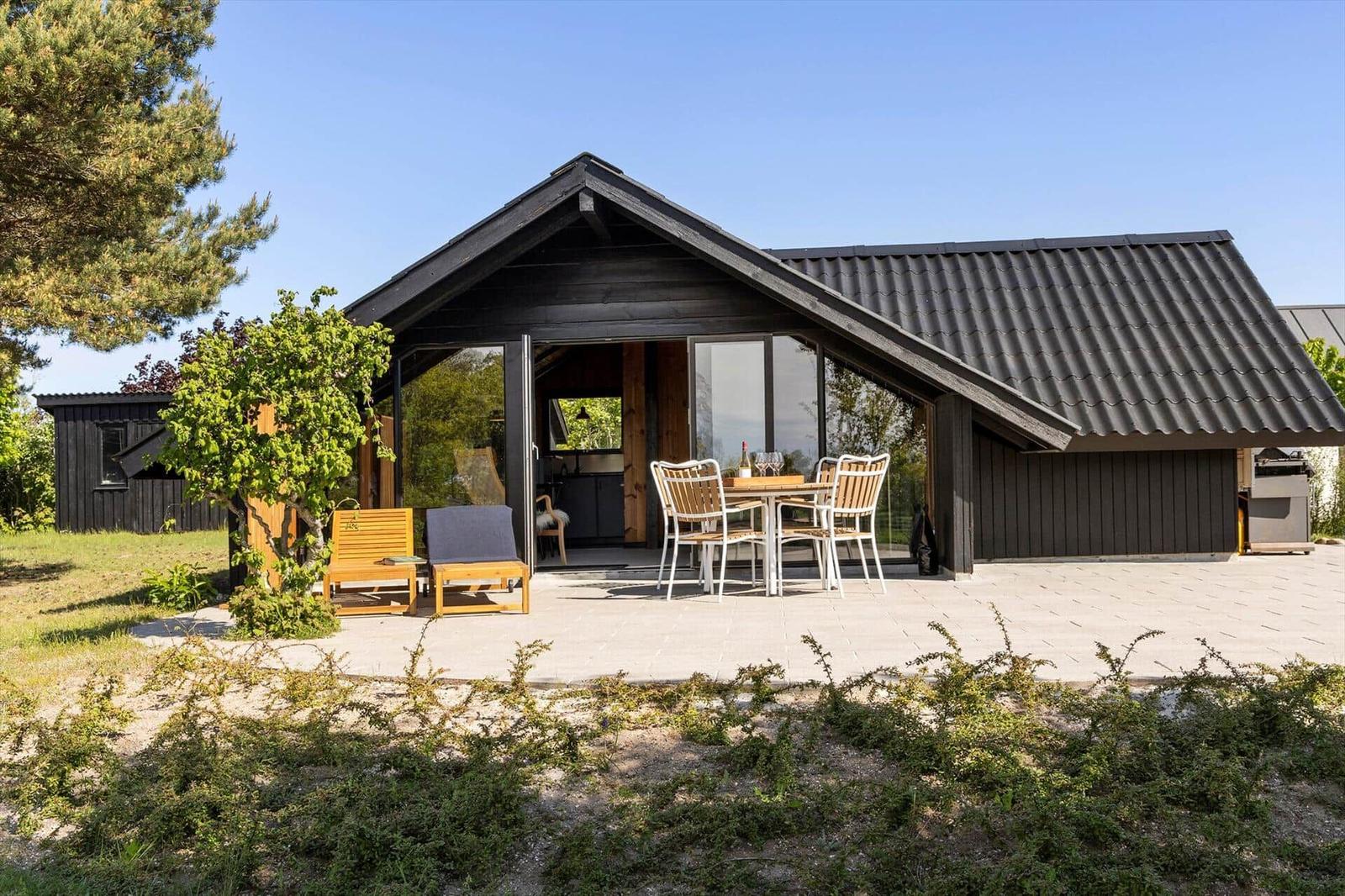 Modern wooden cabin with terrace, table and chairs. Greenery and tree in background.