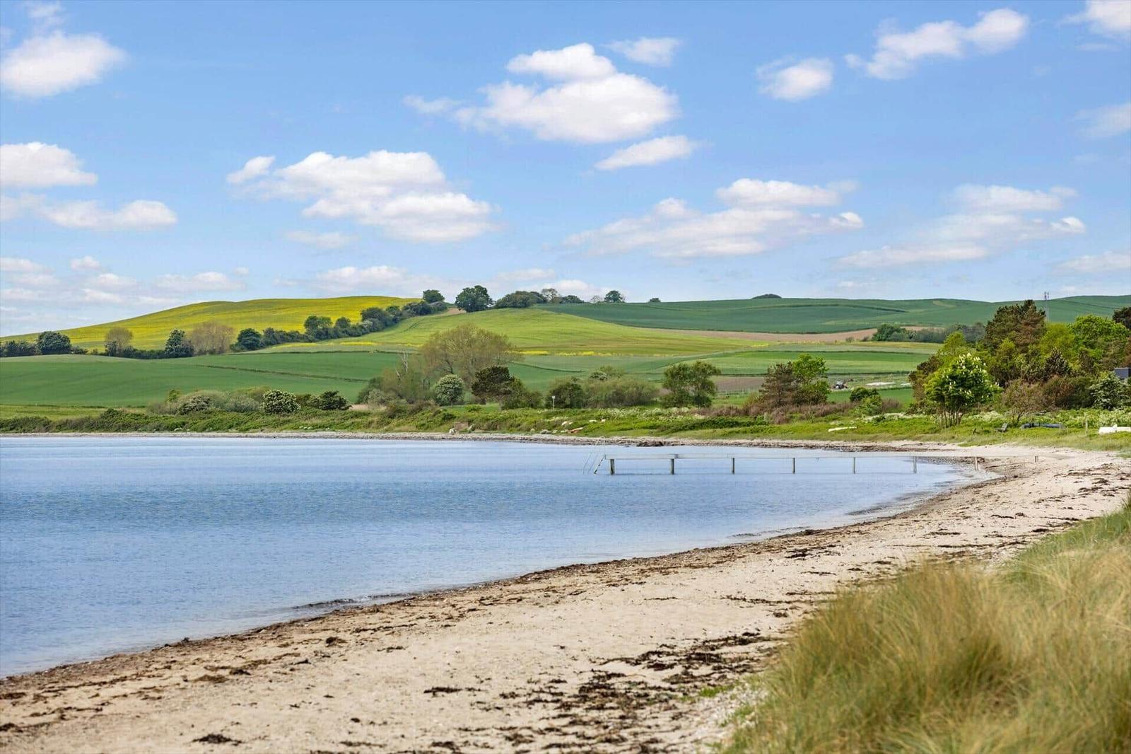 Strand mit Blick auf grüne Hügel und ruhiges Wasser unter blauem Himmel.