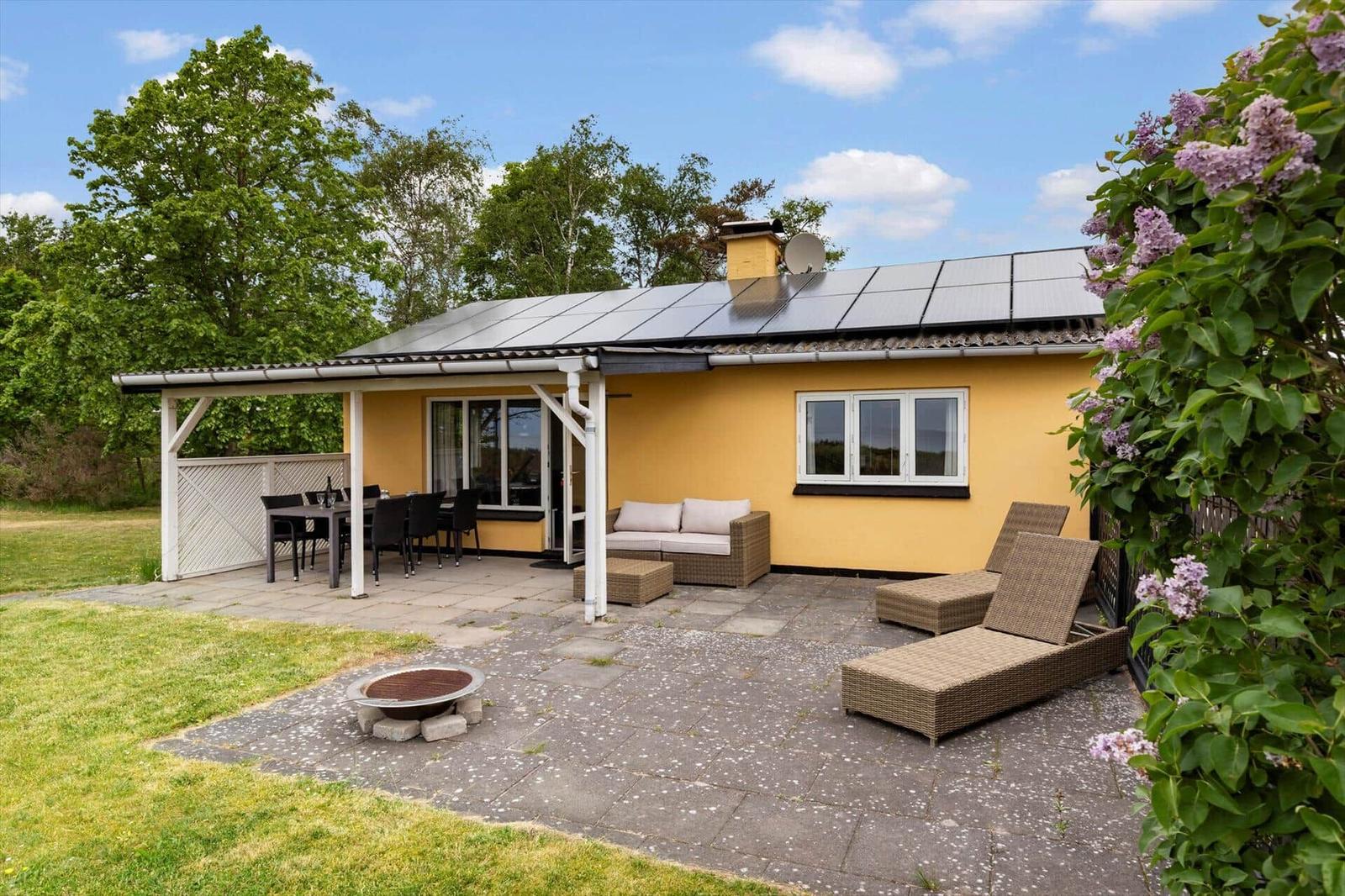 Gelbes Haus mit Terrasse, Sitzgelegenheiten und Solaranlage. Grün umgeben mit Lilien.