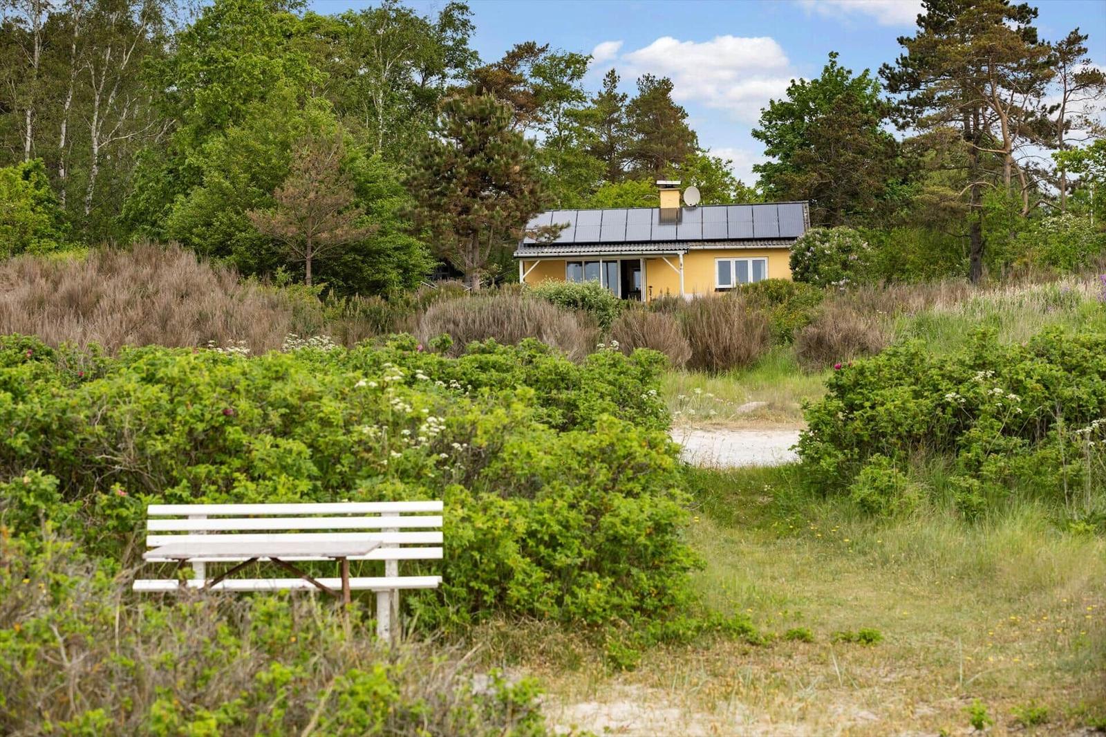Ein gelbes Haus mit Solarpanelen steht inmitten von Grünflächen und Bäumen. Eine weiße Bank ist im Vordergrund.