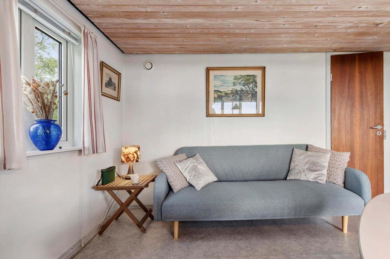 Ein Wohnzimmer mit einem grauen Sofa, einem Holztisch und einem Fenster mit Blumenvase.
