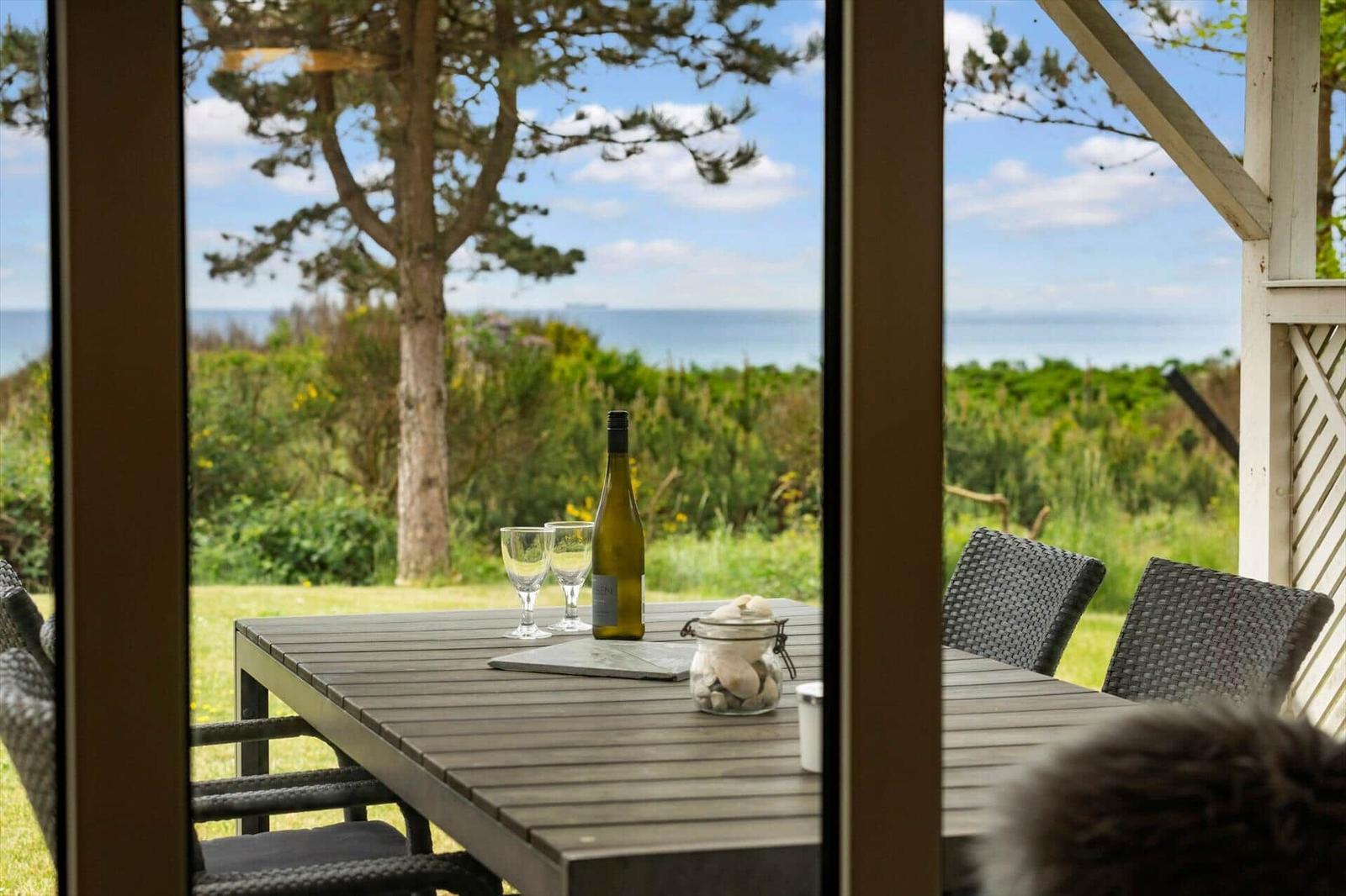 Terrasse mit Tisch, Stühlen, Weinflasche und Blick auf Meer und Baum.