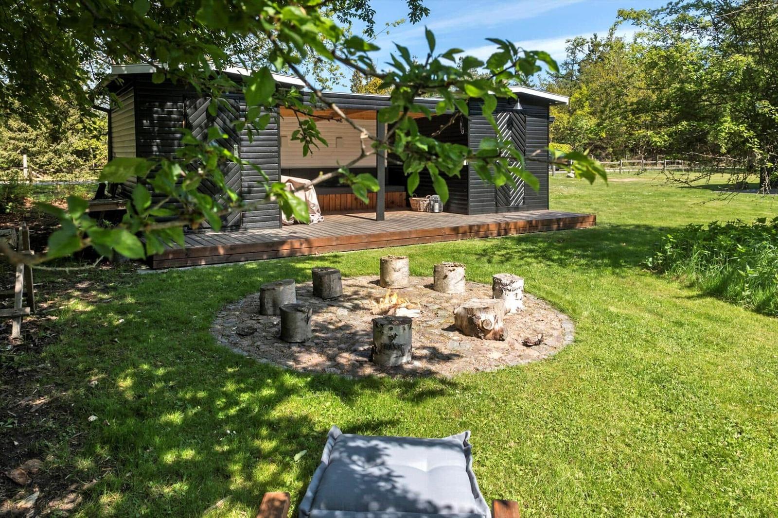 Hütte mit Terrasse und Rundfeuerplatz im Gras. Holzstühle um Feuer.