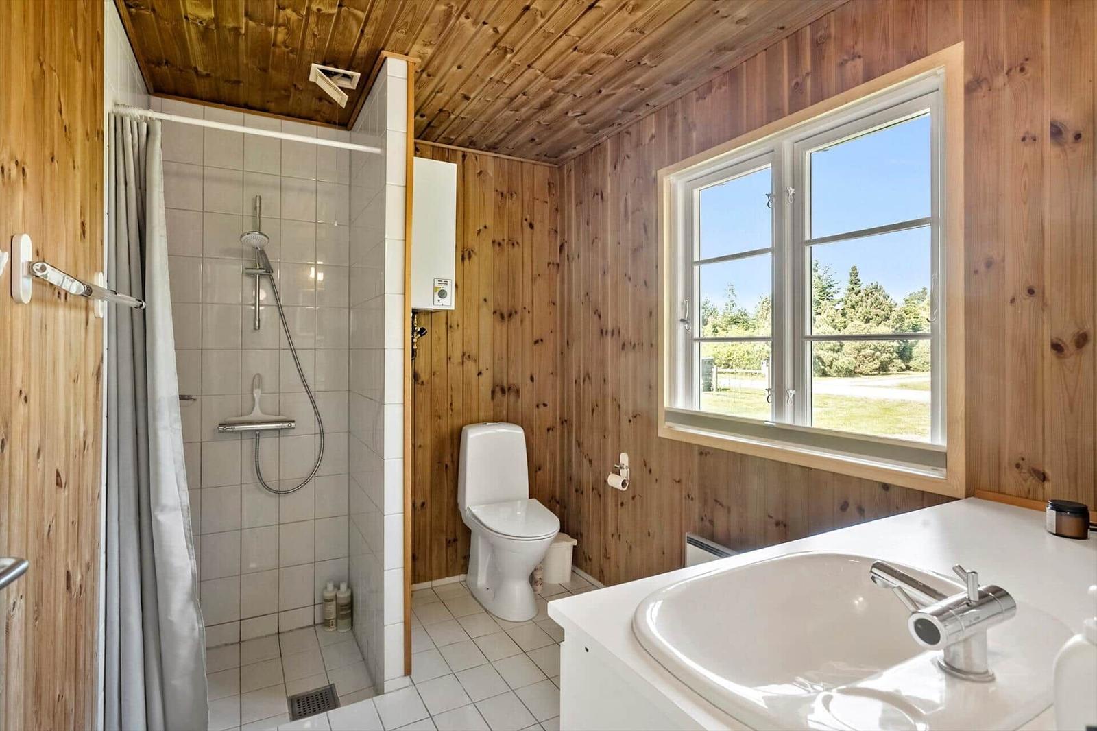Badezimmer mit Dusche, Toilette und Waschbecken. Holzwände und Fenster mit Blick auf Grün.