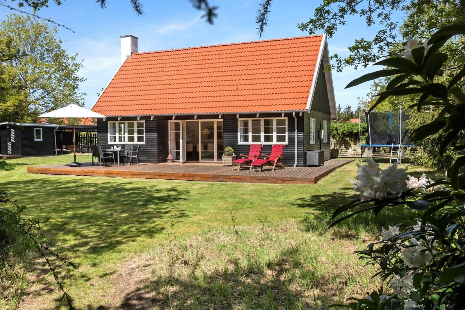 Ein Haus mit rotem Dach und Holzterrasse. Rote Liegestühle und ein Tisch stehen draußen.