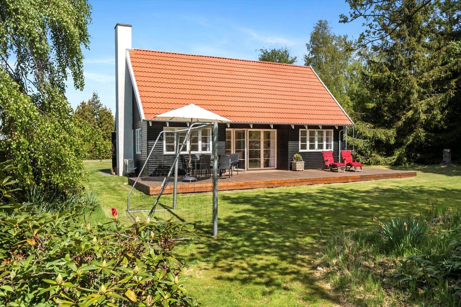 Haus mit Holzterrasse, rotem Dach und Garten. Rote Liegestühle und Sonnenschutz.