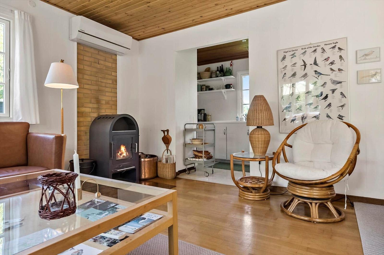 Wohnzimmer mit Kamin, Rattan-Stuhl und Glas-Tisch. Küche im Hintergrund.