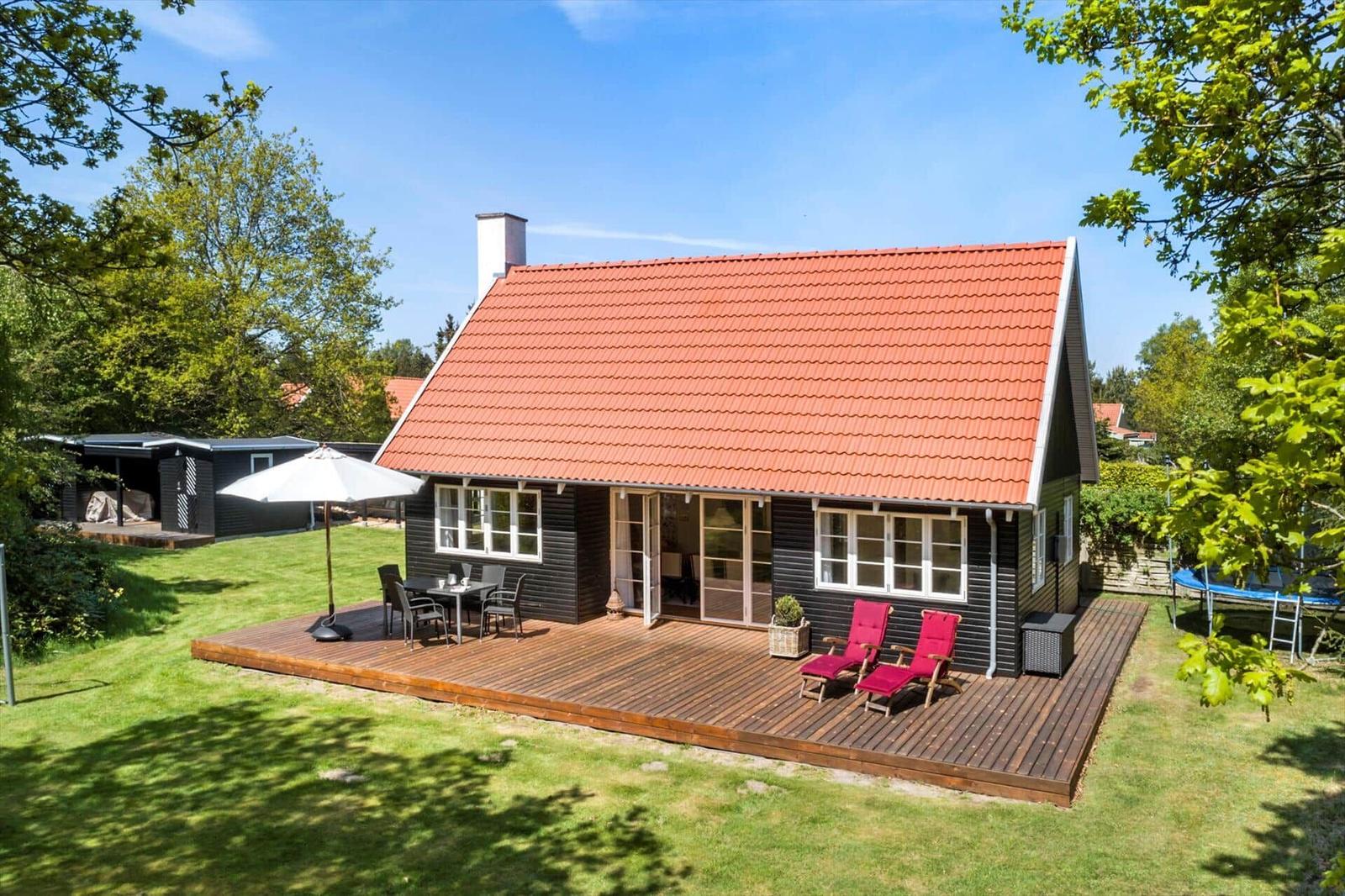 Haus mit Terrasse, Garten und rotem Dach. Rote Liegestühle und Tisch unter Sonnenschirm.
