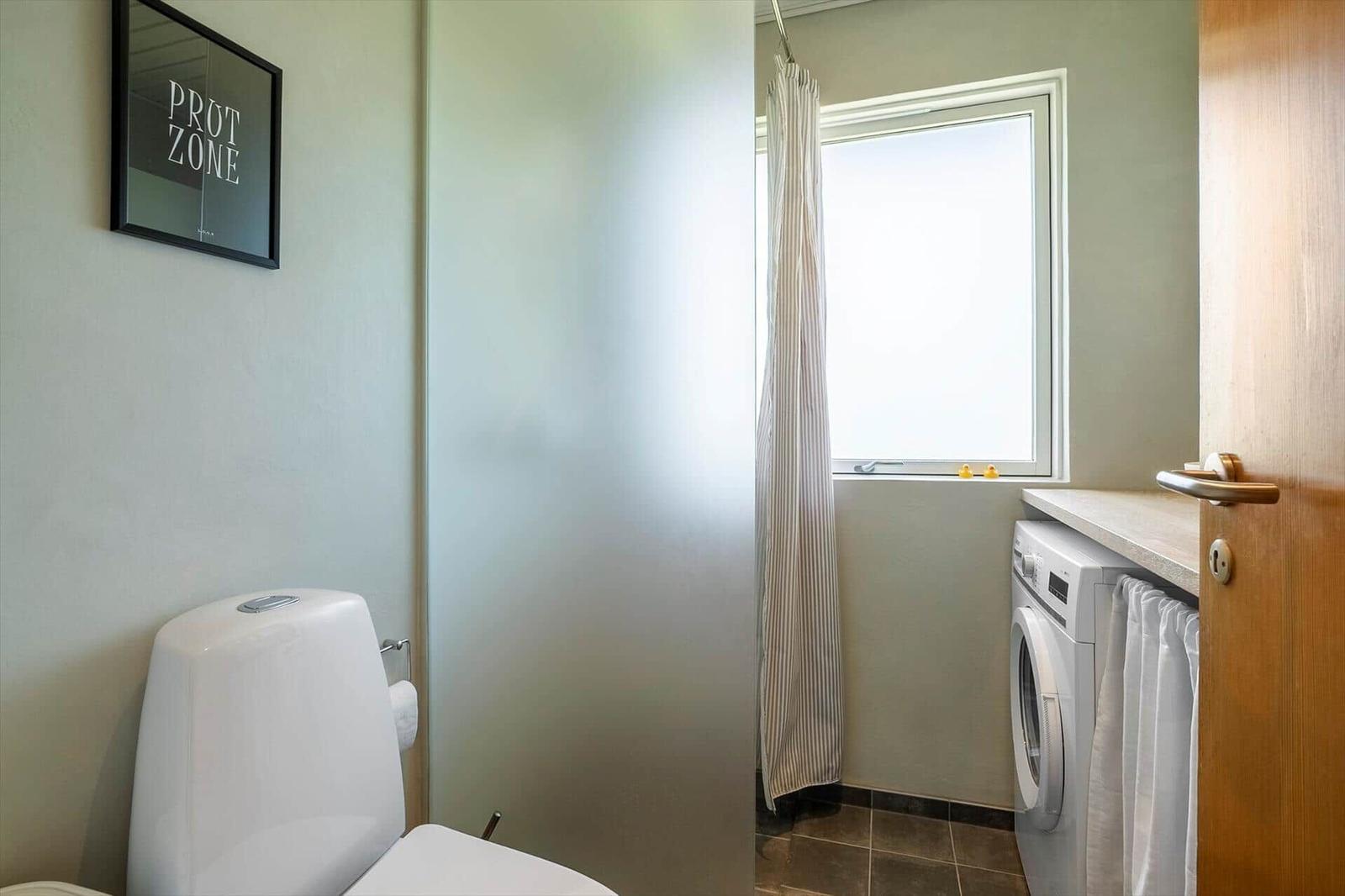 Bathroom with toilet, washing machine, and window with curtain.