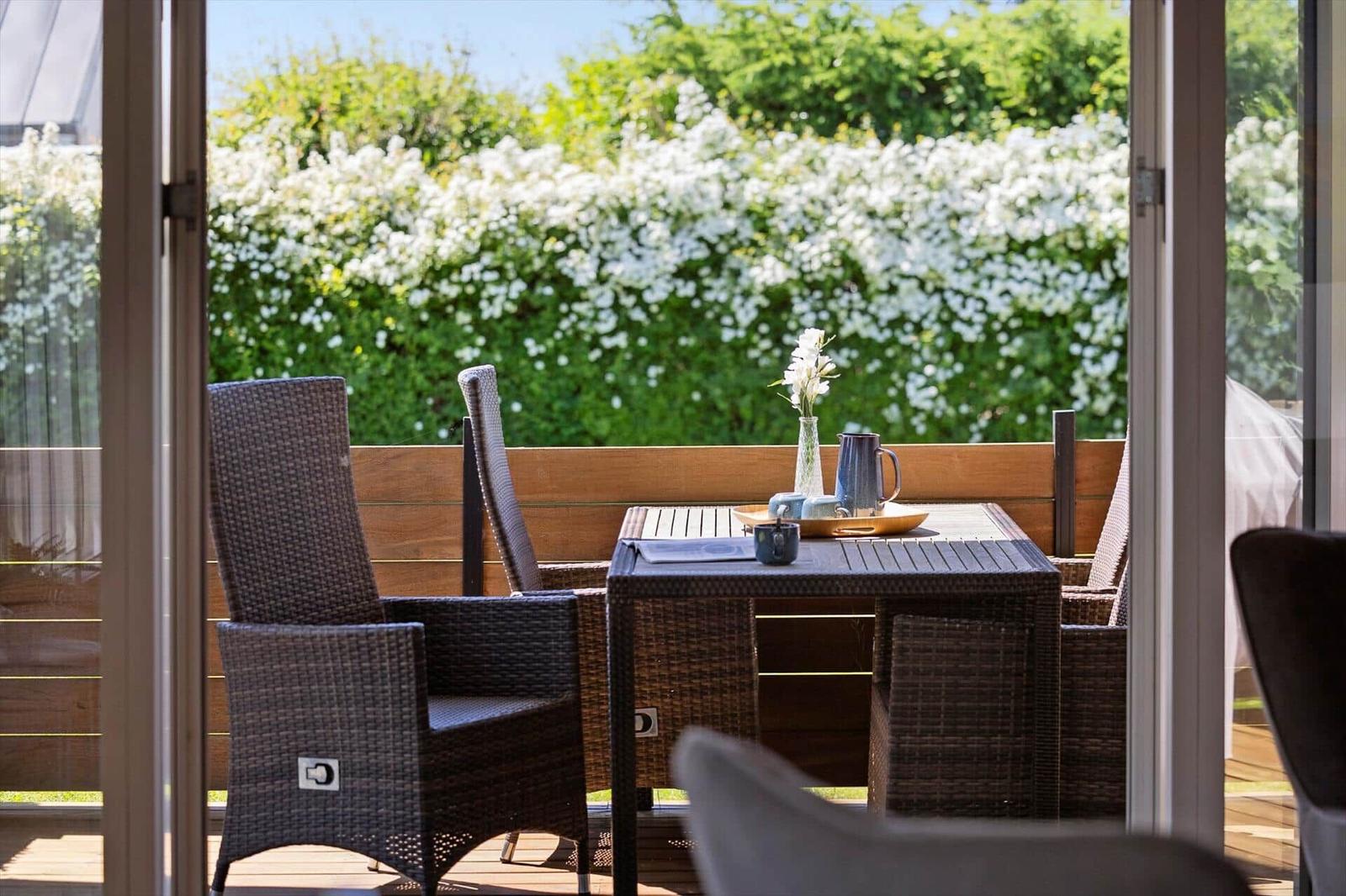 Terrace with table and chairs, view of green hedge with white flowers.