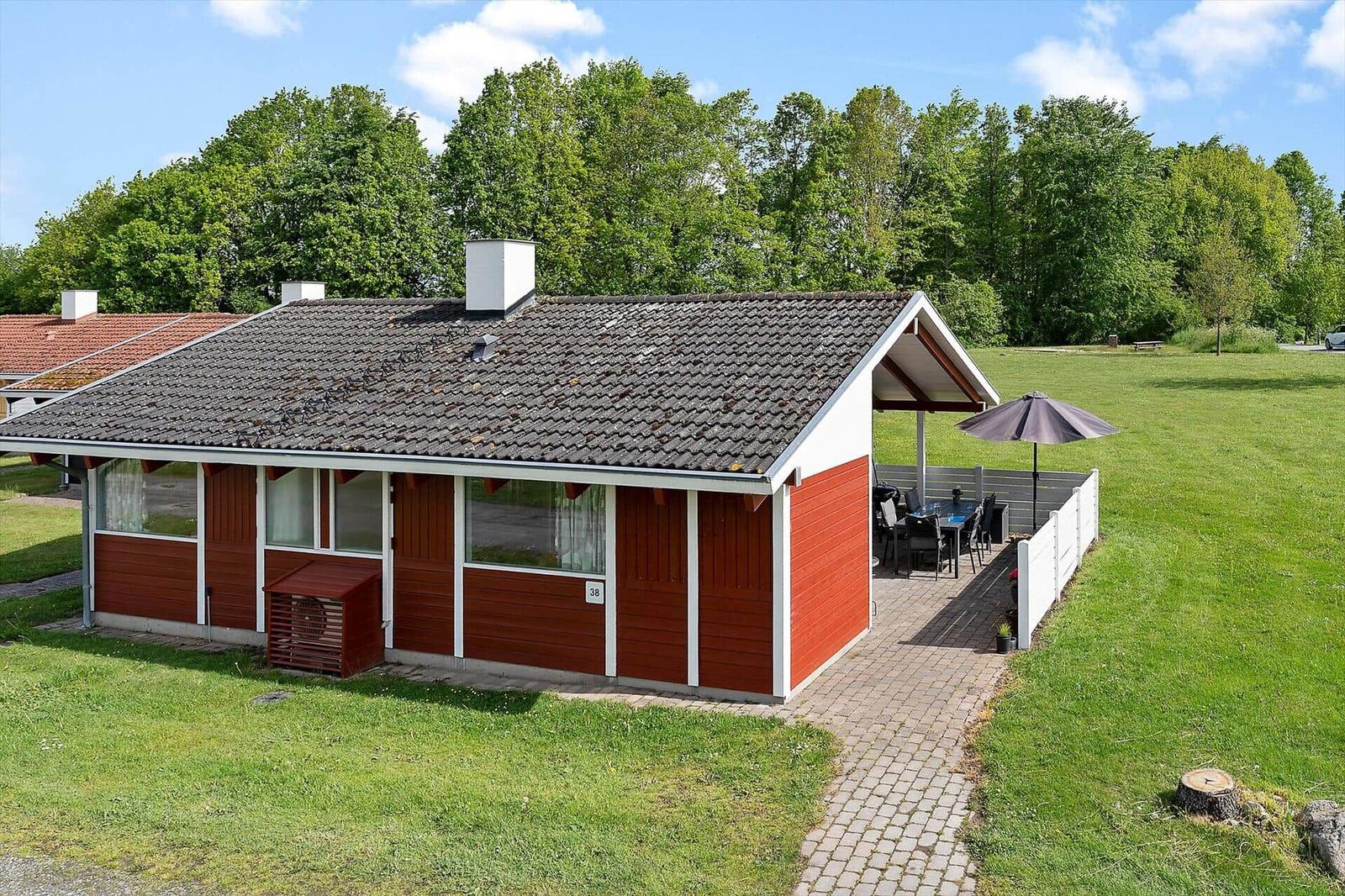 Red holiday home with terrace, umbrella, and garden. Background: green trees.
