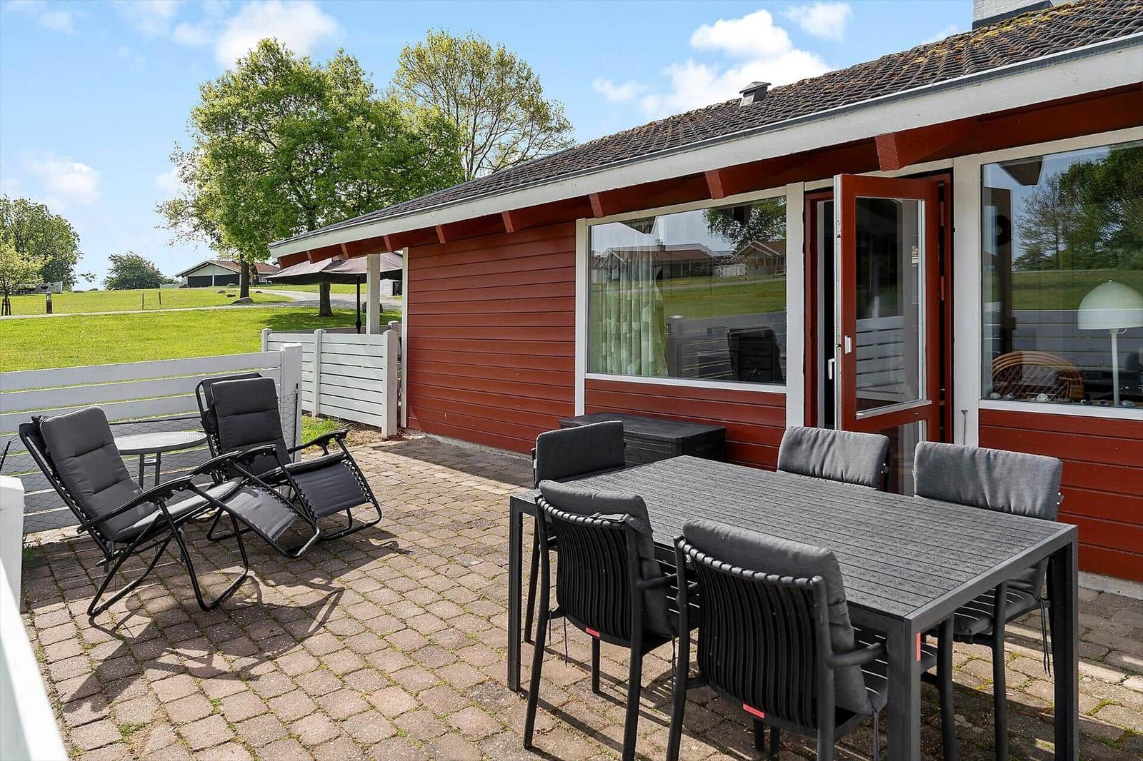 Terrace with table, chairs, and lounge chairs in front of a red house with a white fence.
