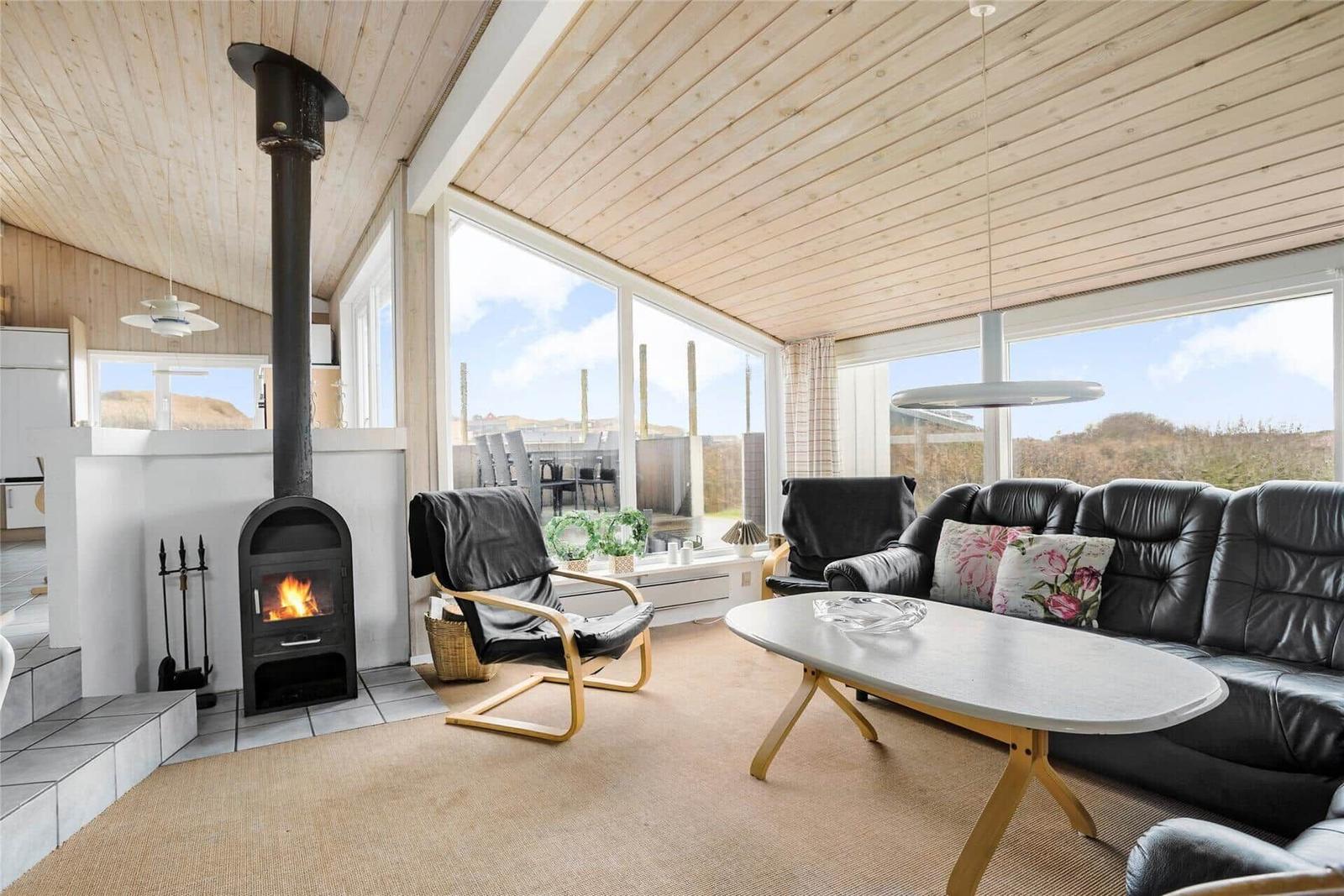 Wohnzimmer mit Kamin, Sofa und Sessel. Große Fenster bieten Blick auf die Landschaft.