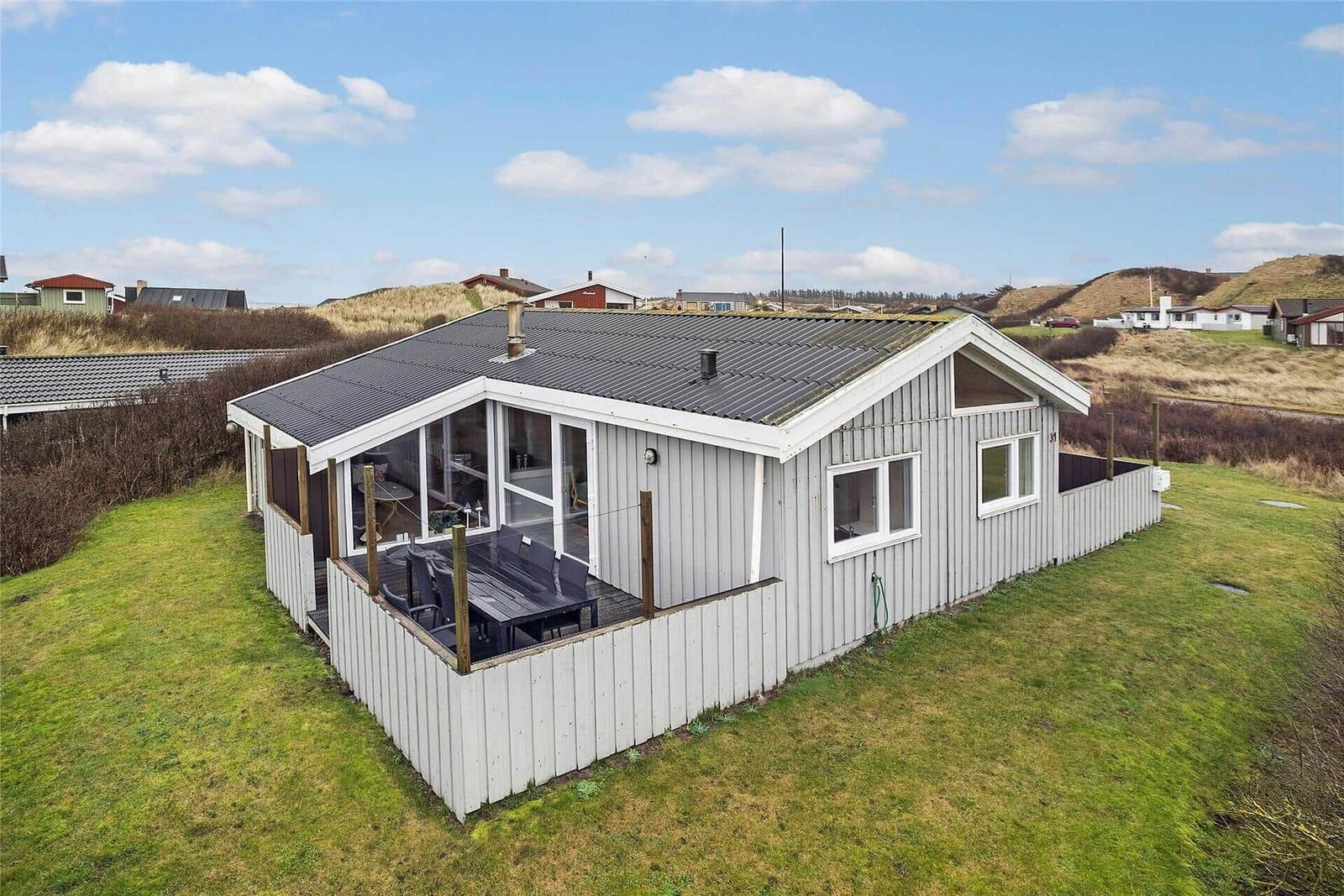 Ein Ferienhaus mit Terrasse und Garten. Im Hintergrund sind Hügel und andere Häuser sichtbar.