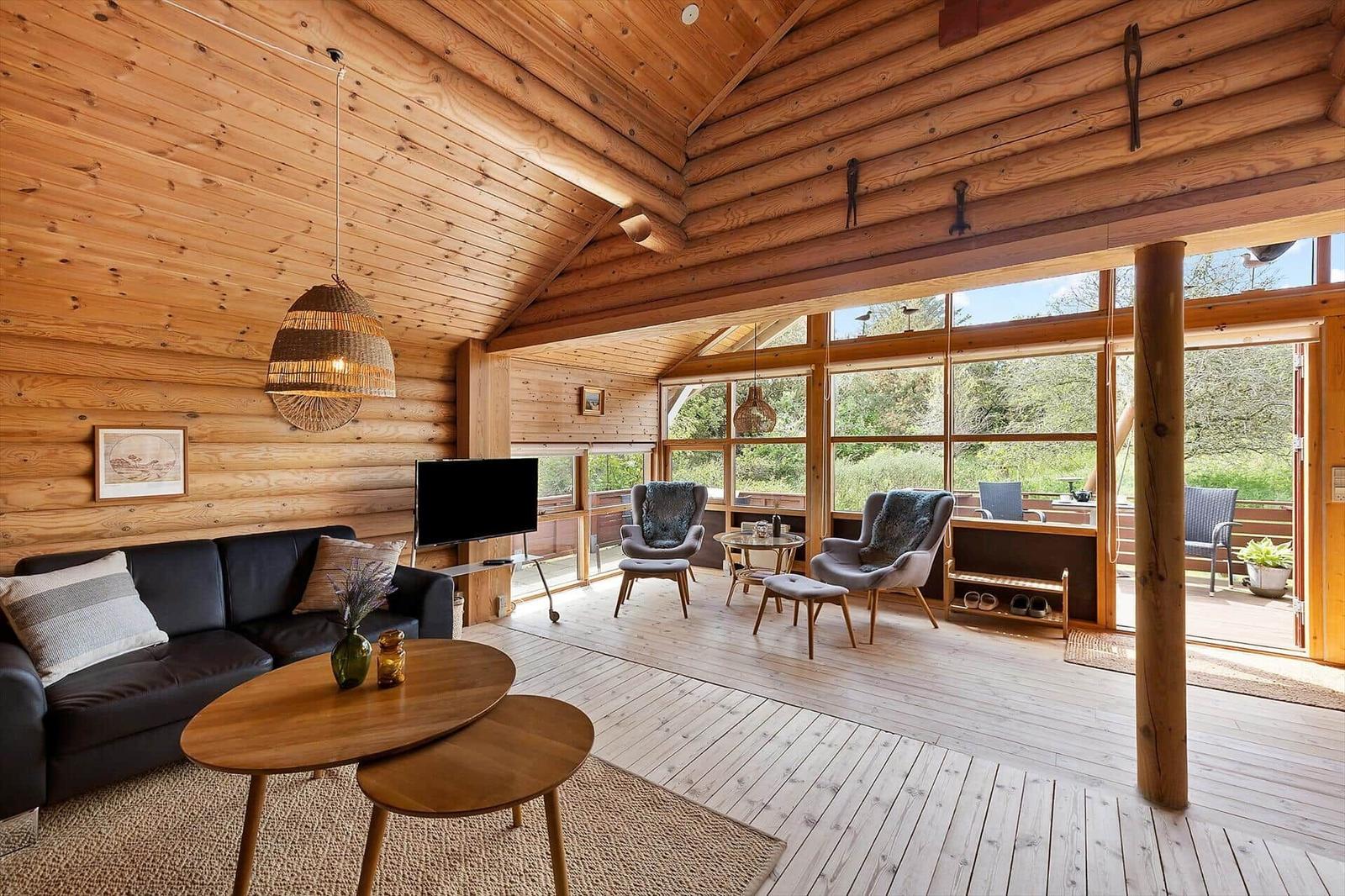 Wohnzimmer mit Holzwand, Sofa, Fernseher und Sitzgelegenheiten. Große Fenster mit Blick nach draußen.