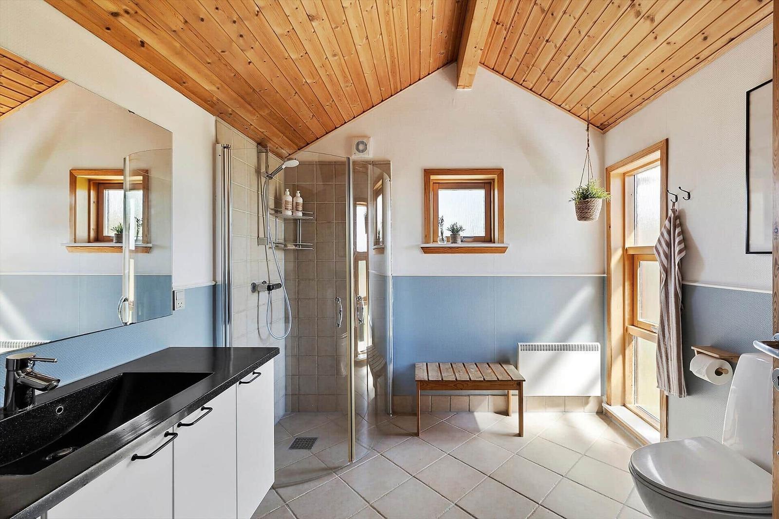 Badezimmer mit Dusche, Waschbecken und Toilette. Holzdecke und Fenster mit Holzrahmen.
