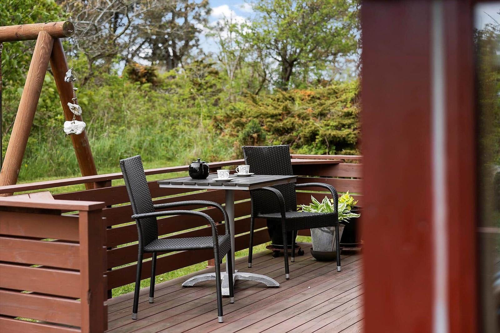 Terrasse mit Tisch, zwei Stühlen und Pflanzen. Hintergrund: Grünflächen und Bäume.