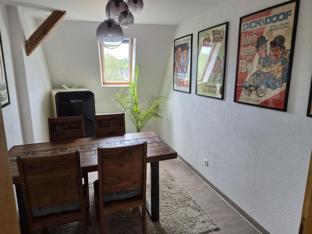 Esszimmer mit Holztisch, Stühlen, Kühlschrank und Wandplakaten.