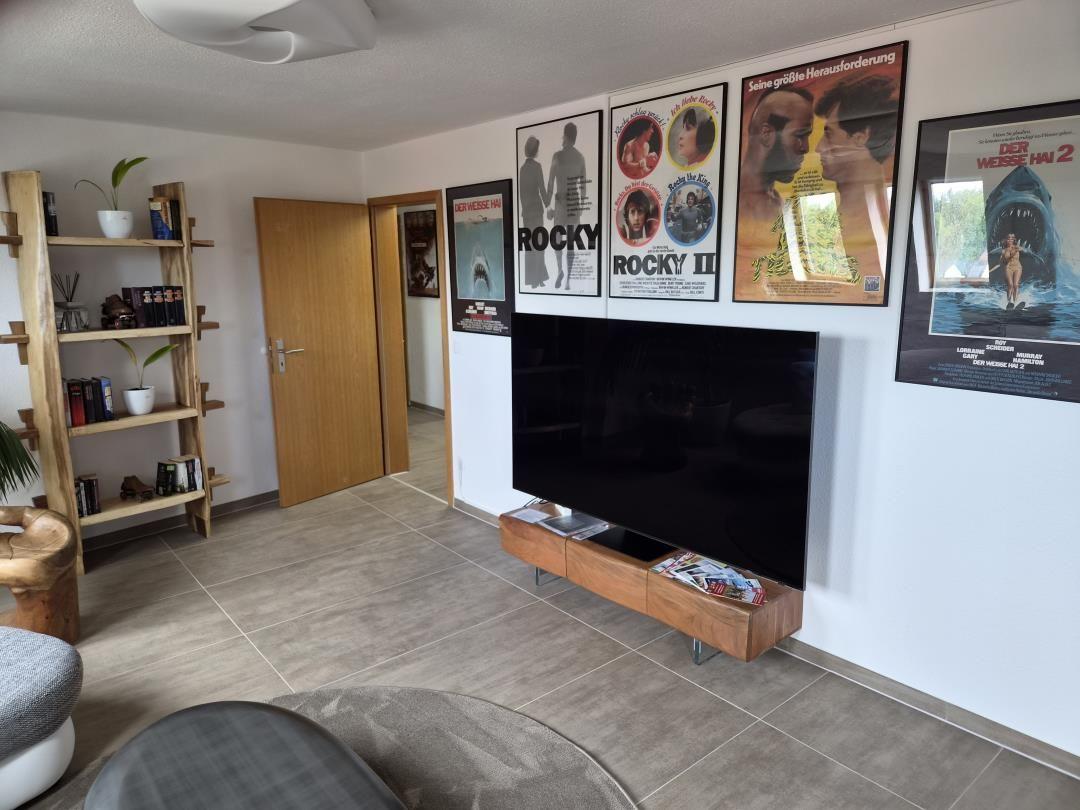 Wohnzimmer mit Fernseher, Filmplakaten und Holzregal. Fliesenboden und Tür im Hintergrund.